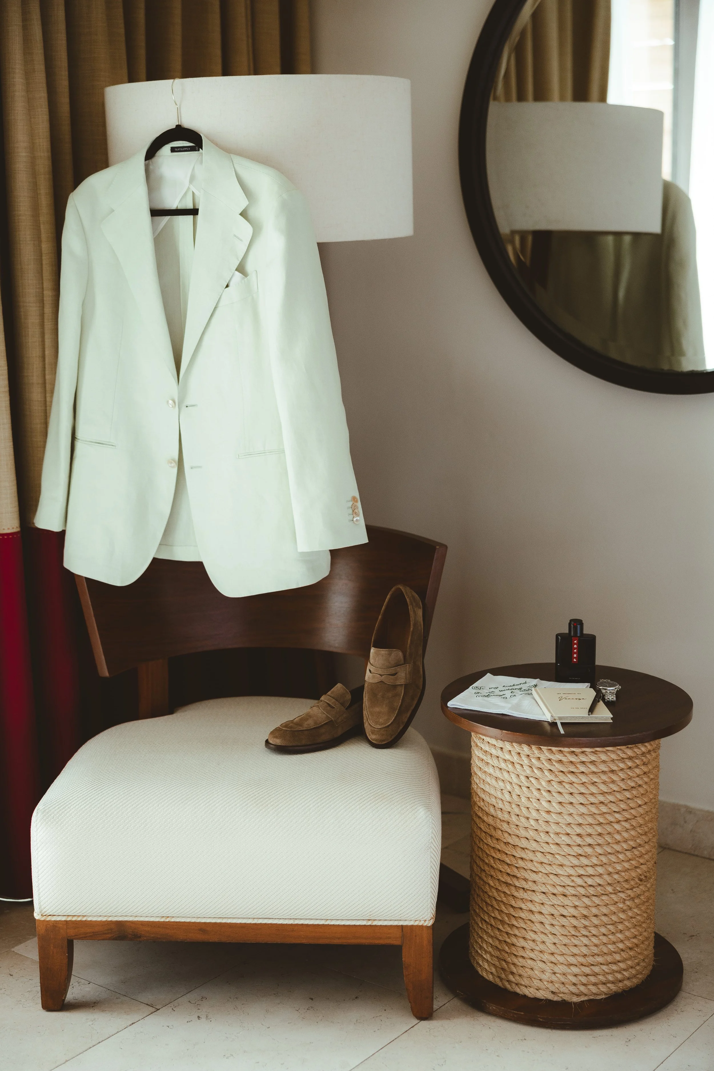 A white blazer hanging on a hanger on a lamp, shoes on a chair, and books and personal items on a side table next to a beige upholstered chair, with a round mirror on the wall and curtains in the background. It's the groom detail's at Secrets Akumal 