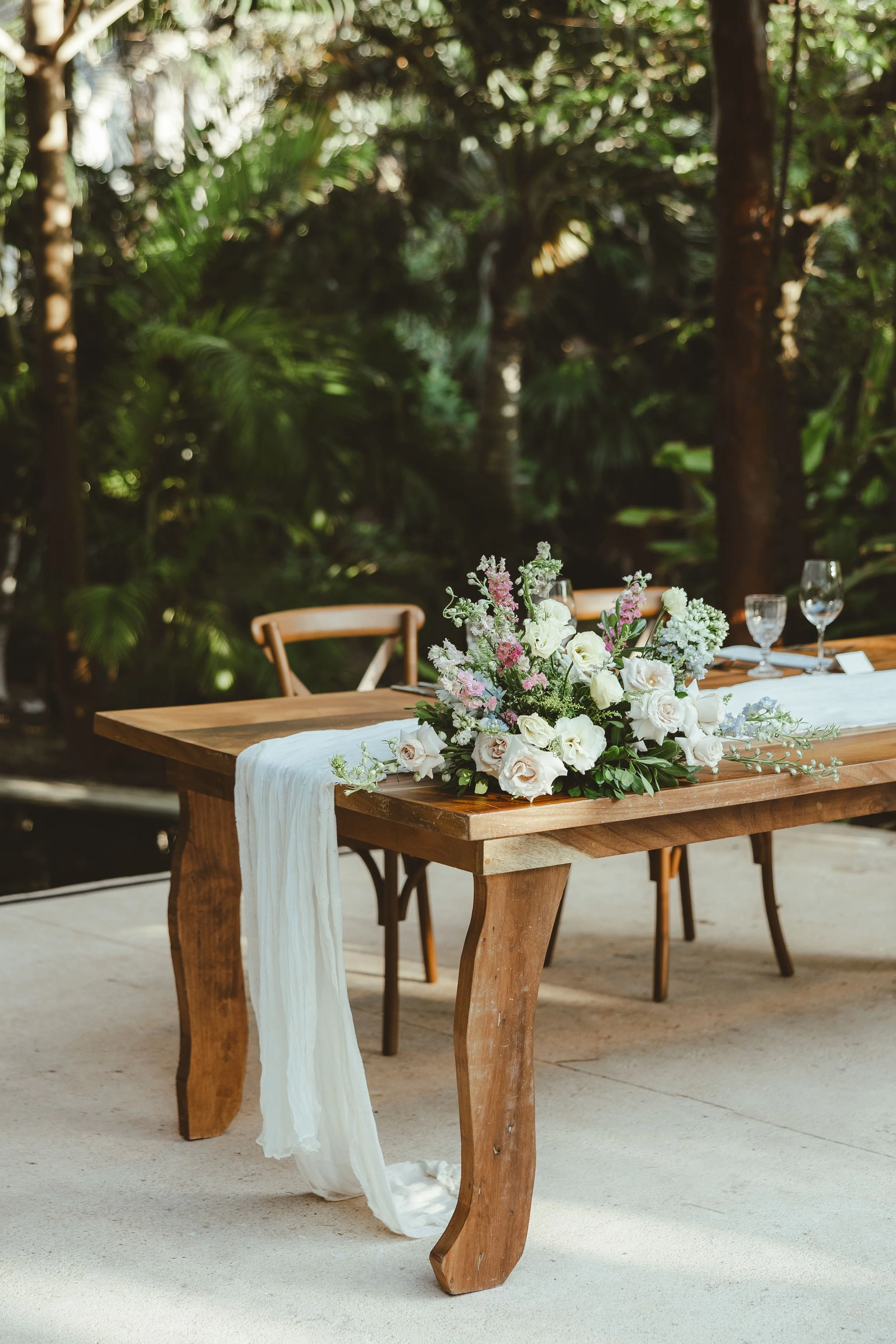 A wooden table decorated with a floral centerpiece, white cloth, and glassware set up outdoors in a lush green garden.