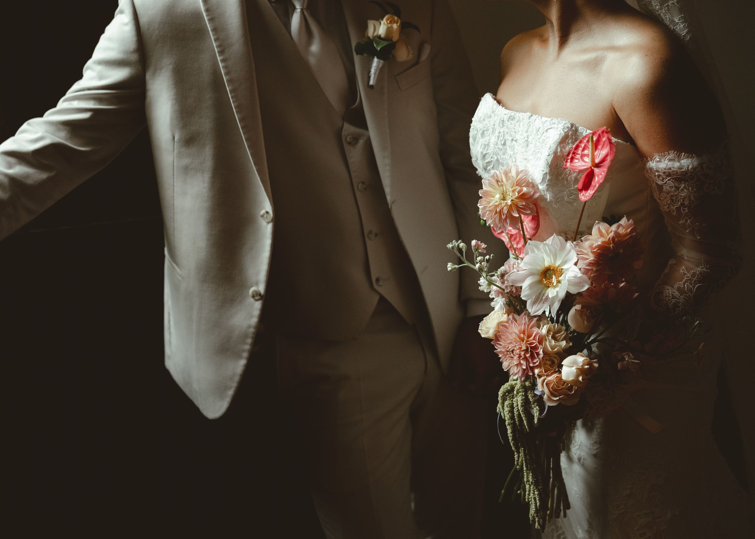 A bride in a white wedding dress holding a bouquet of pink, white, and peach flowers, standing next to a groom in a light gray suit with a boutonniere of white flowers and dark green leaves.