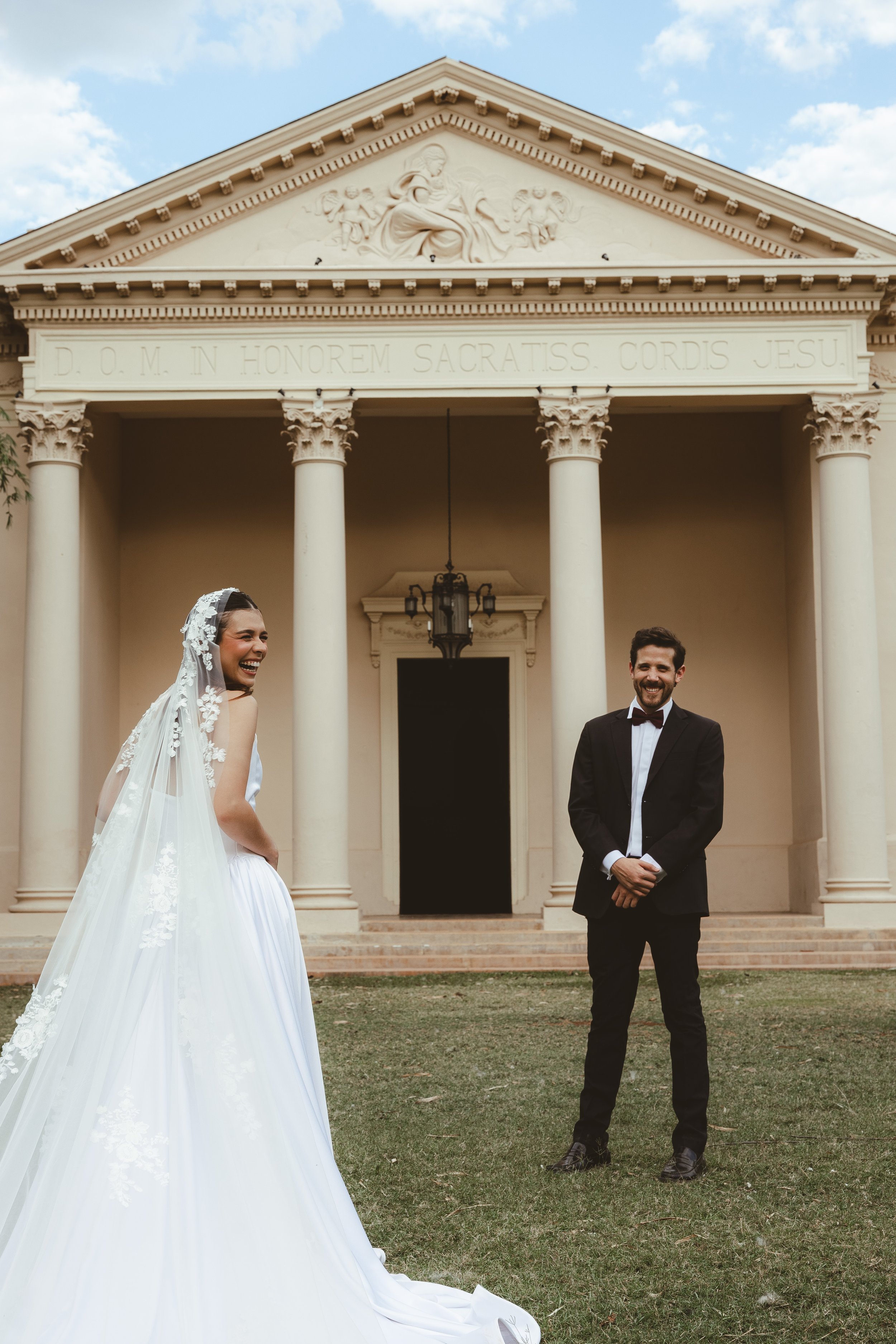 A bride and groom standing in front of a classical building with columns, smiling and looking at each other. The bride is wearing a white wedding gown with lace details and a veil, while the groom is dressed in a black tuxedo with a bow tie.