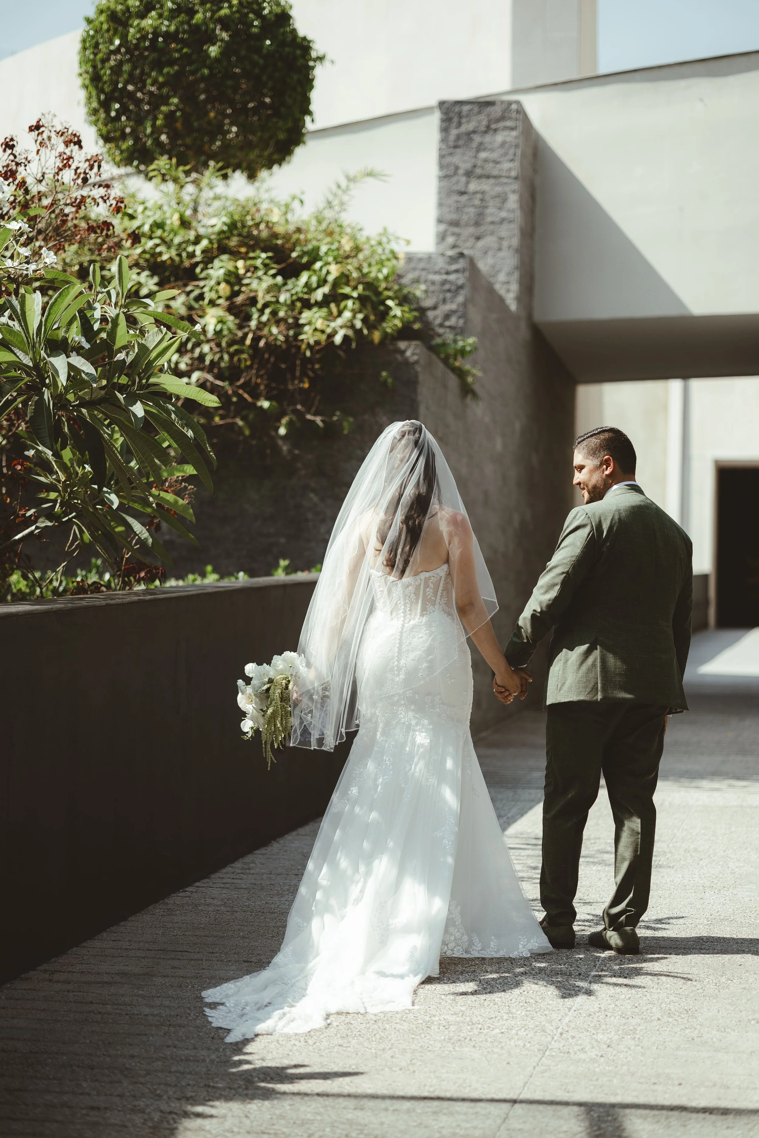 A bride in a white wedding dress with a sheer veil and a groom in a grey suit walking hand in hand outdoors on a sunny day, with modern buildings and greenery around.