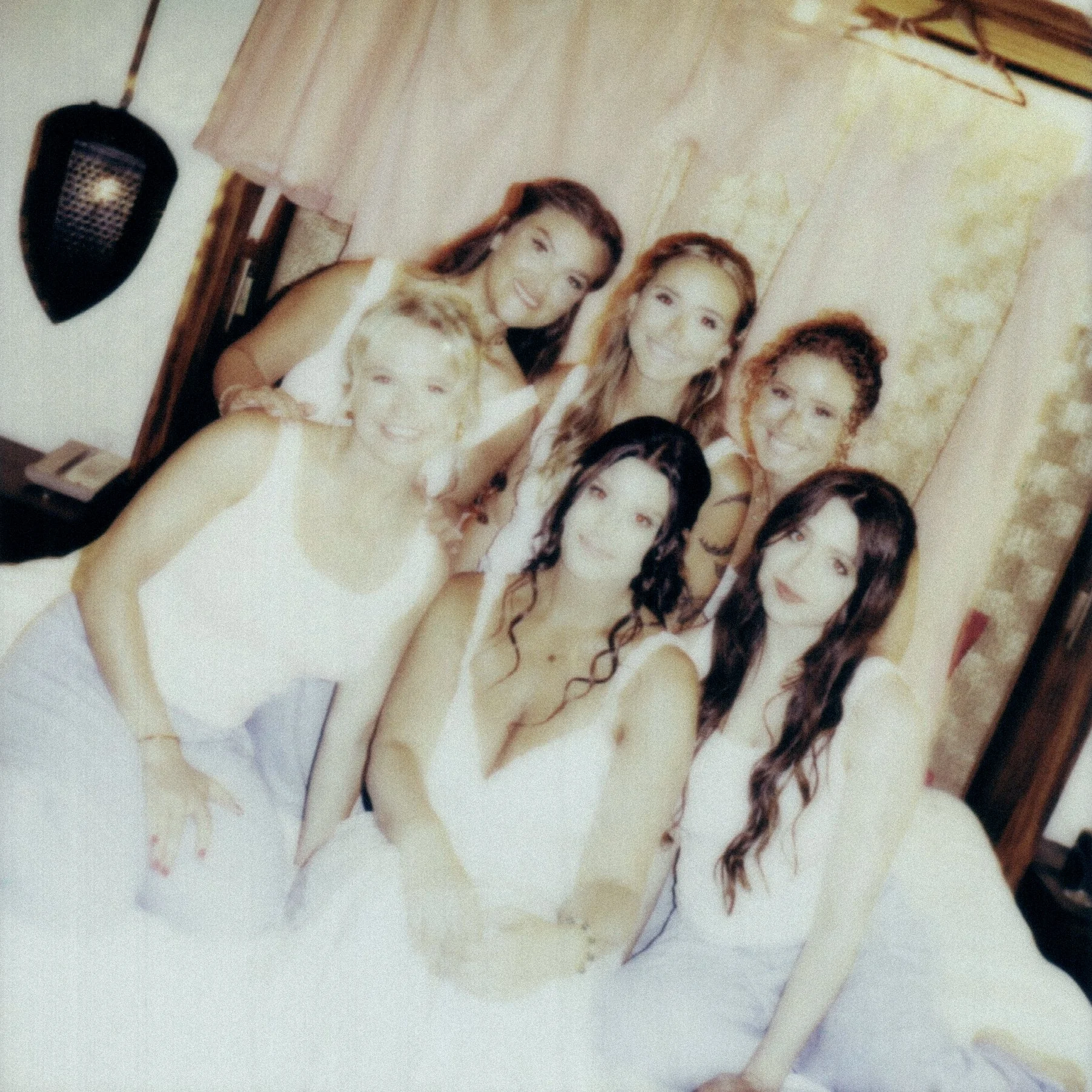 A group of women dressed in white dresses at a wedding, standing close together, smiling for a photo inside a room with beige walls and hanging panels.