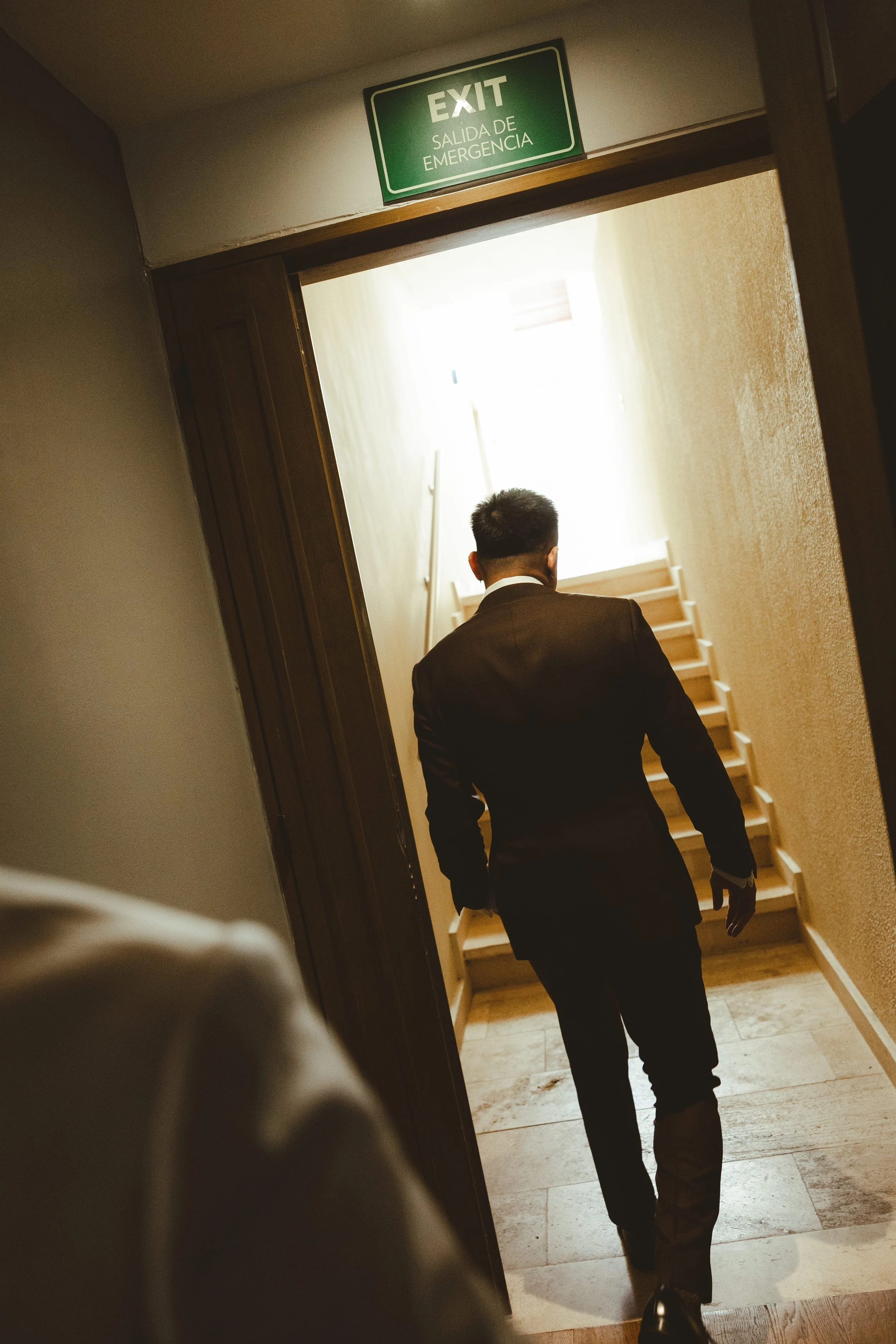 A man in a dark suit walking up a wooden staircase, viewed from behind, with an emergency exit sign above the doorway.