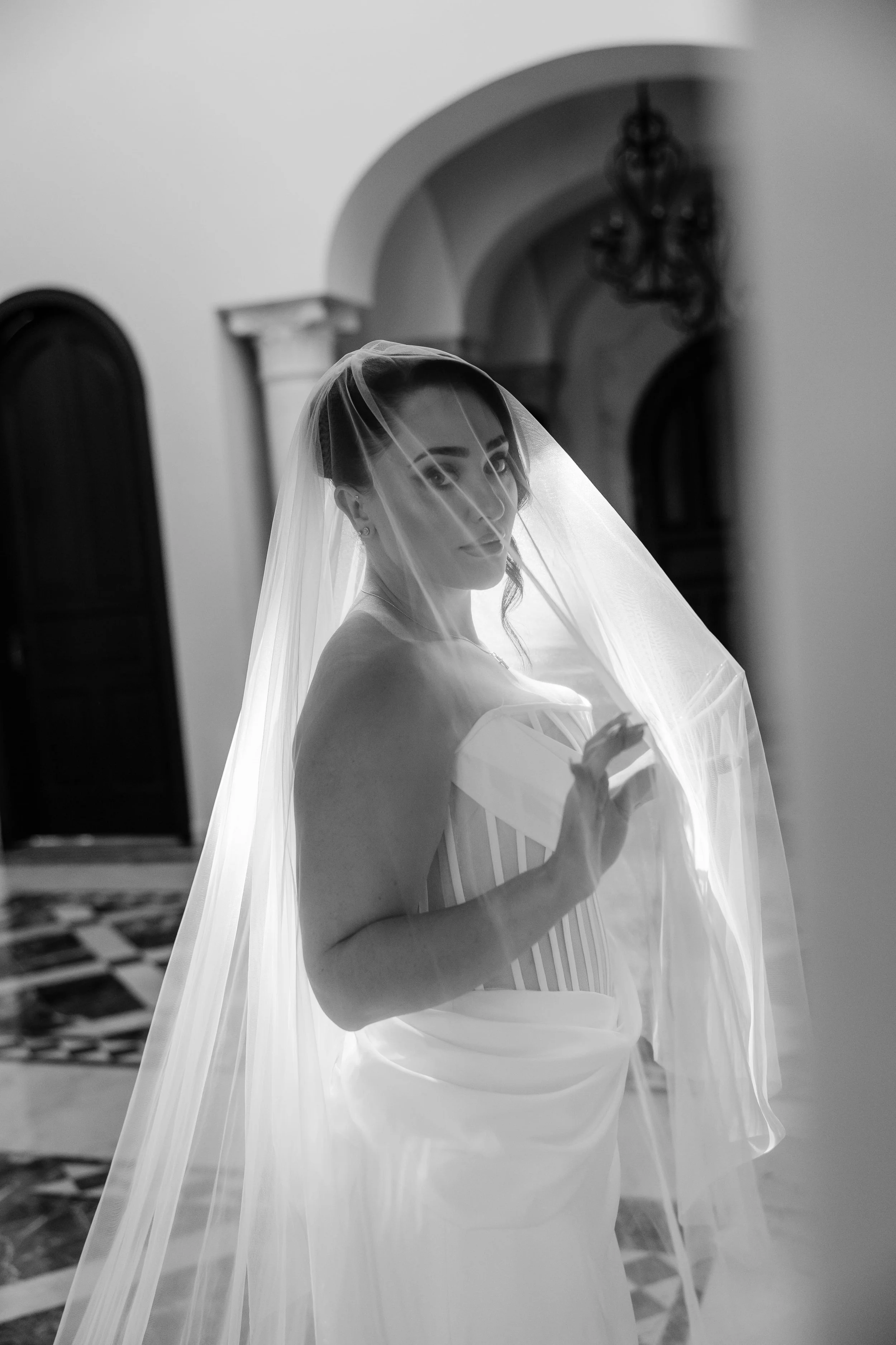 A bride with dark hair styled in an updo, wearing dangling earrings and a strapless wedding dress, stands indoors behind a veil.