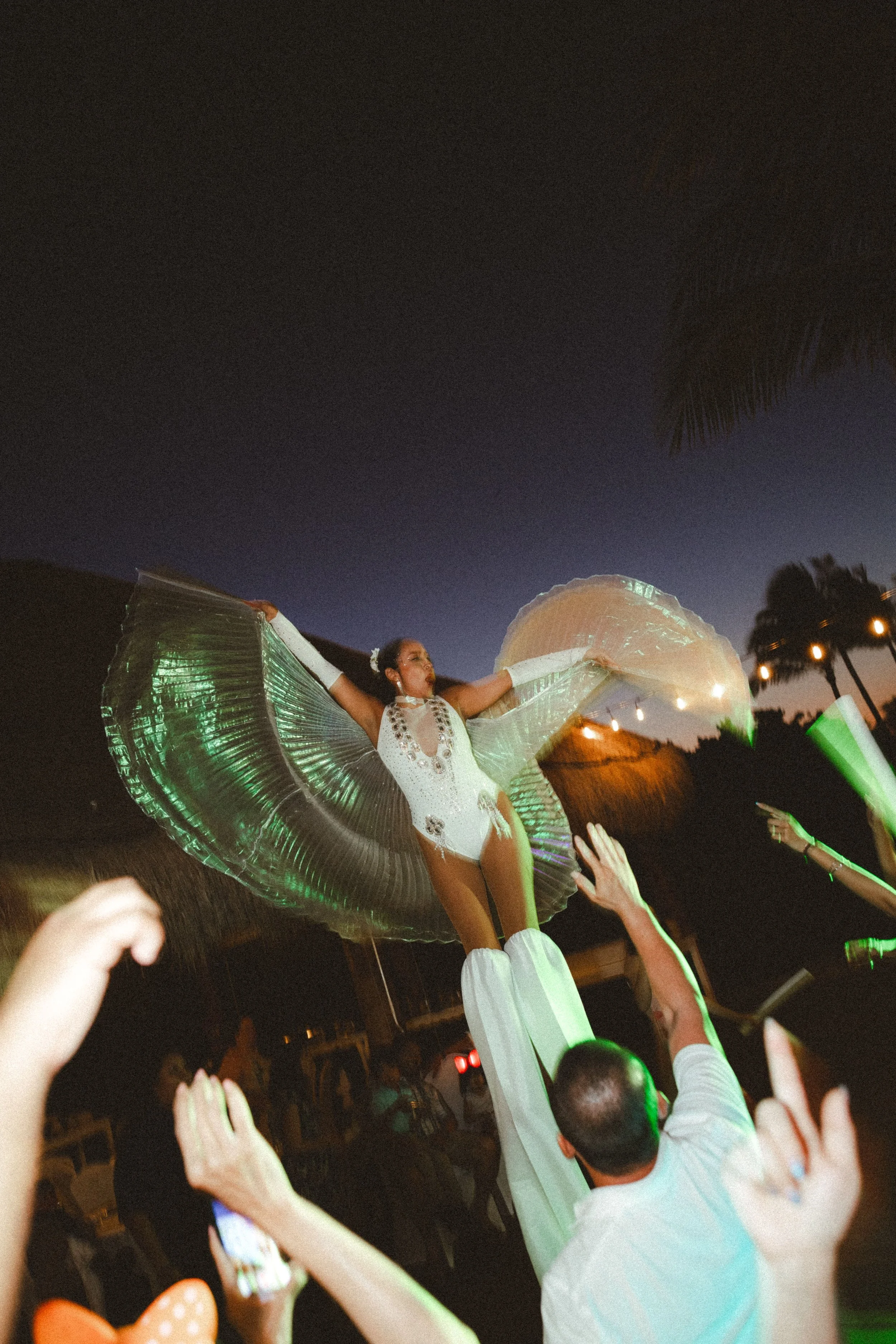 A performer in a white dress with beads is holding a large illuminated parasol above her head while dancing at a nighttime outdoor event. She is surrounded by an audience with raised hands, some taking photos or videos. The background shows a dark sk