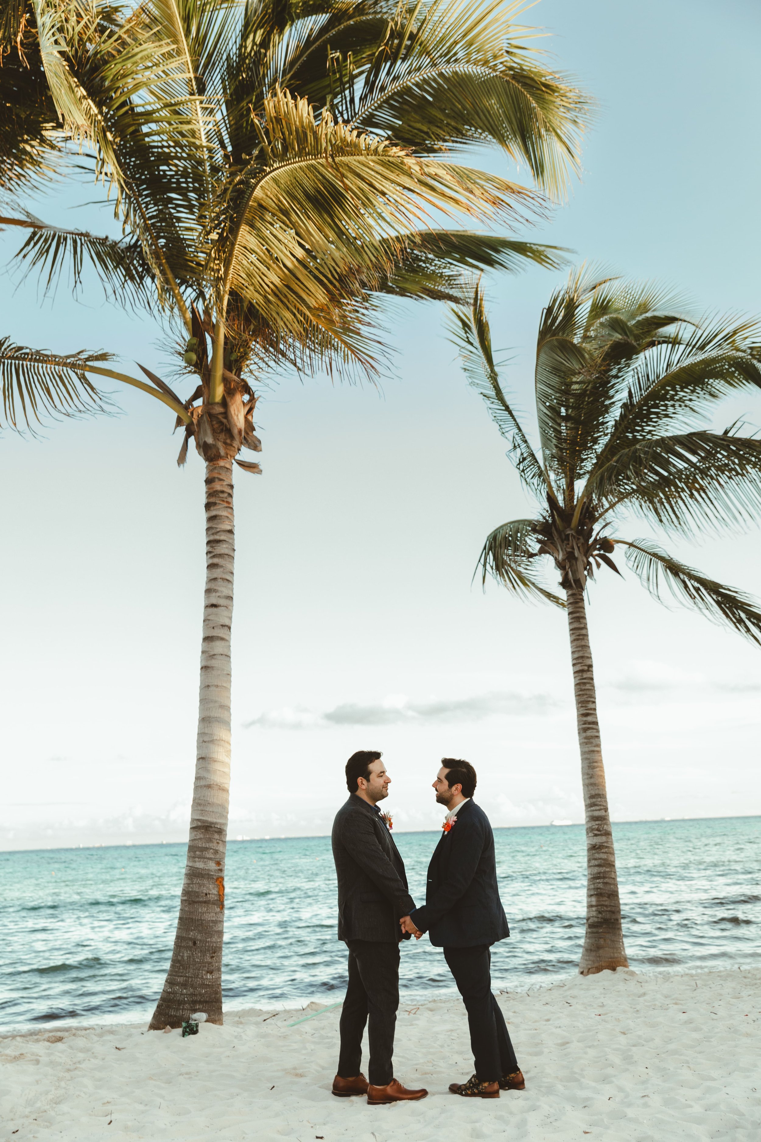Two grooms in suits holding hands and facing each other on a beach with palm trees and ocean in the background, during a wedding their wedding ceremony in Tulum.