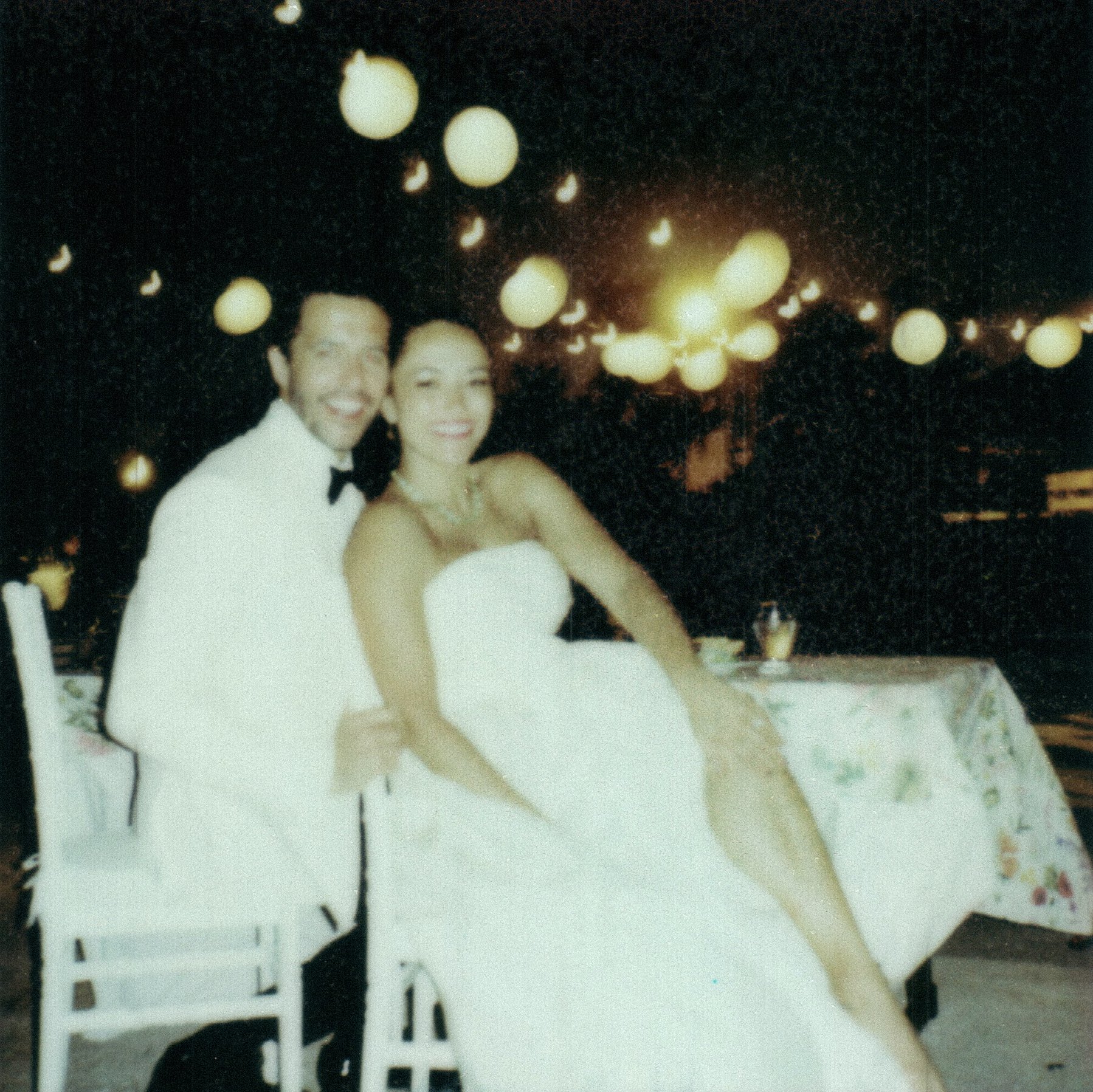 A bride and groom dressed in wedding attire, smiling for a photo at night with a black background and hanging glowing lanterns.