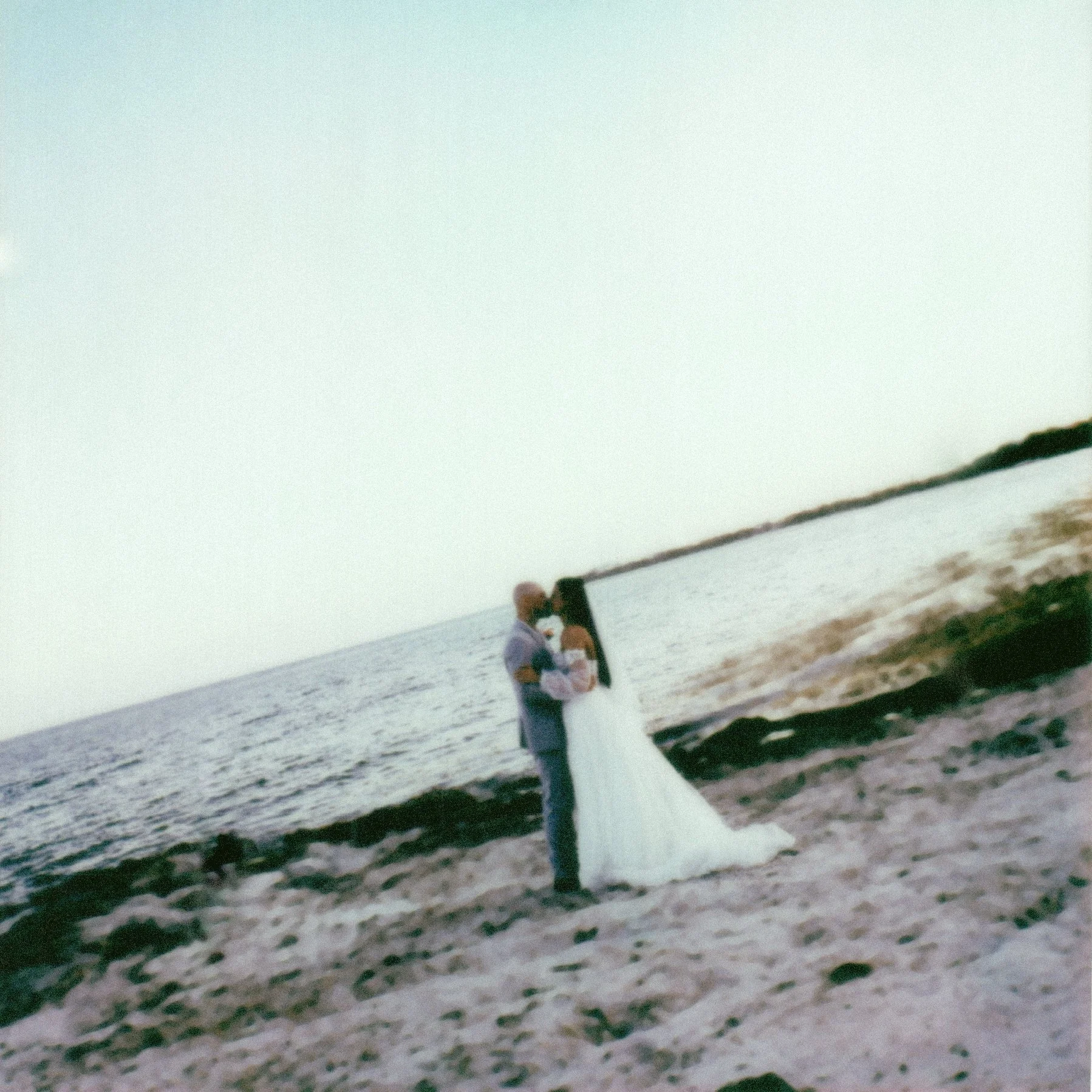 A person sitting on a white bench at the beach, facing the ocean, with a pole in front of them and a small tree or bush in the foreground on a sandy shore.
