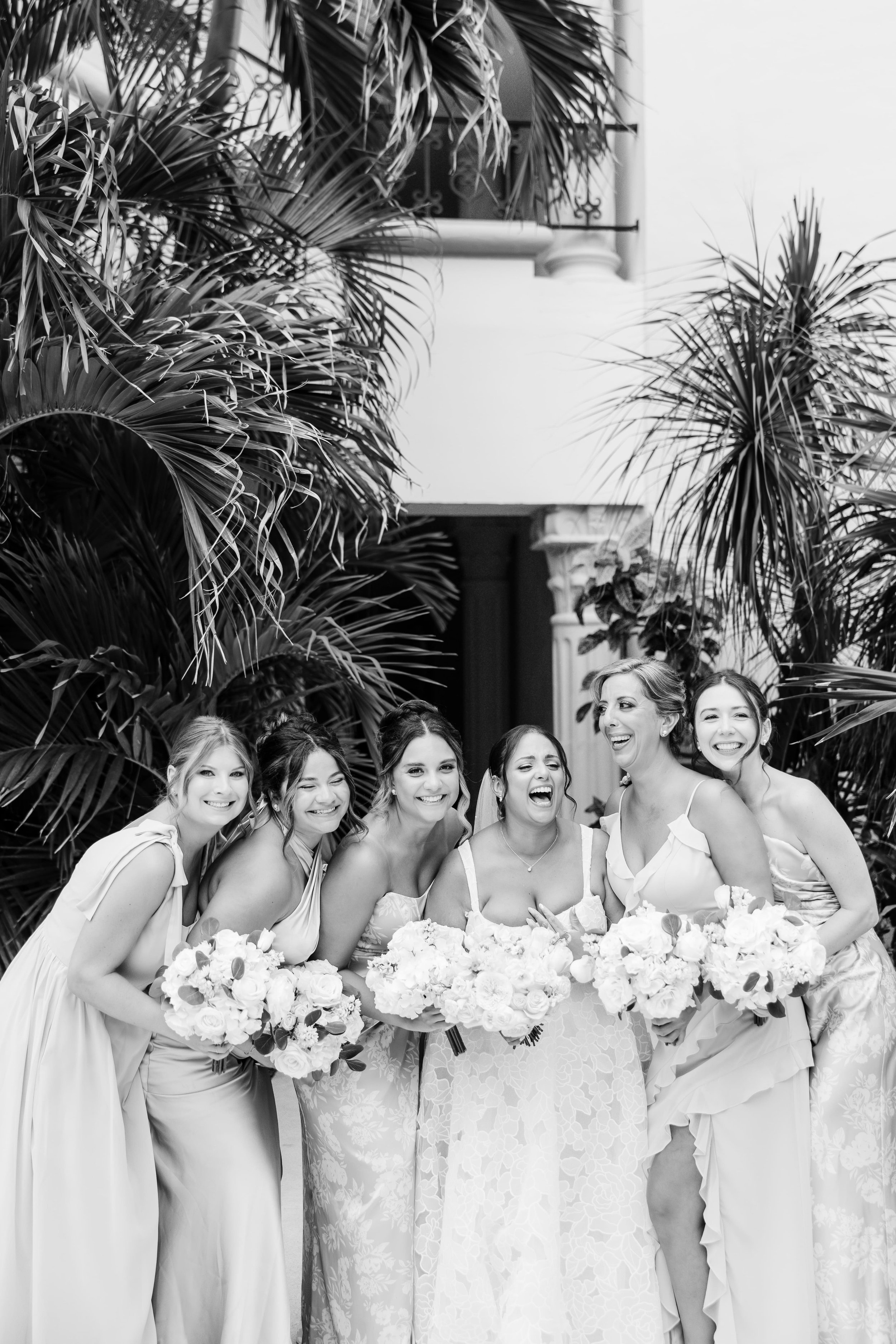 A group of women, including a bride in a lace dress, smiling and posing together at a wedding reception, surrounded by large tropical plants and greenery.