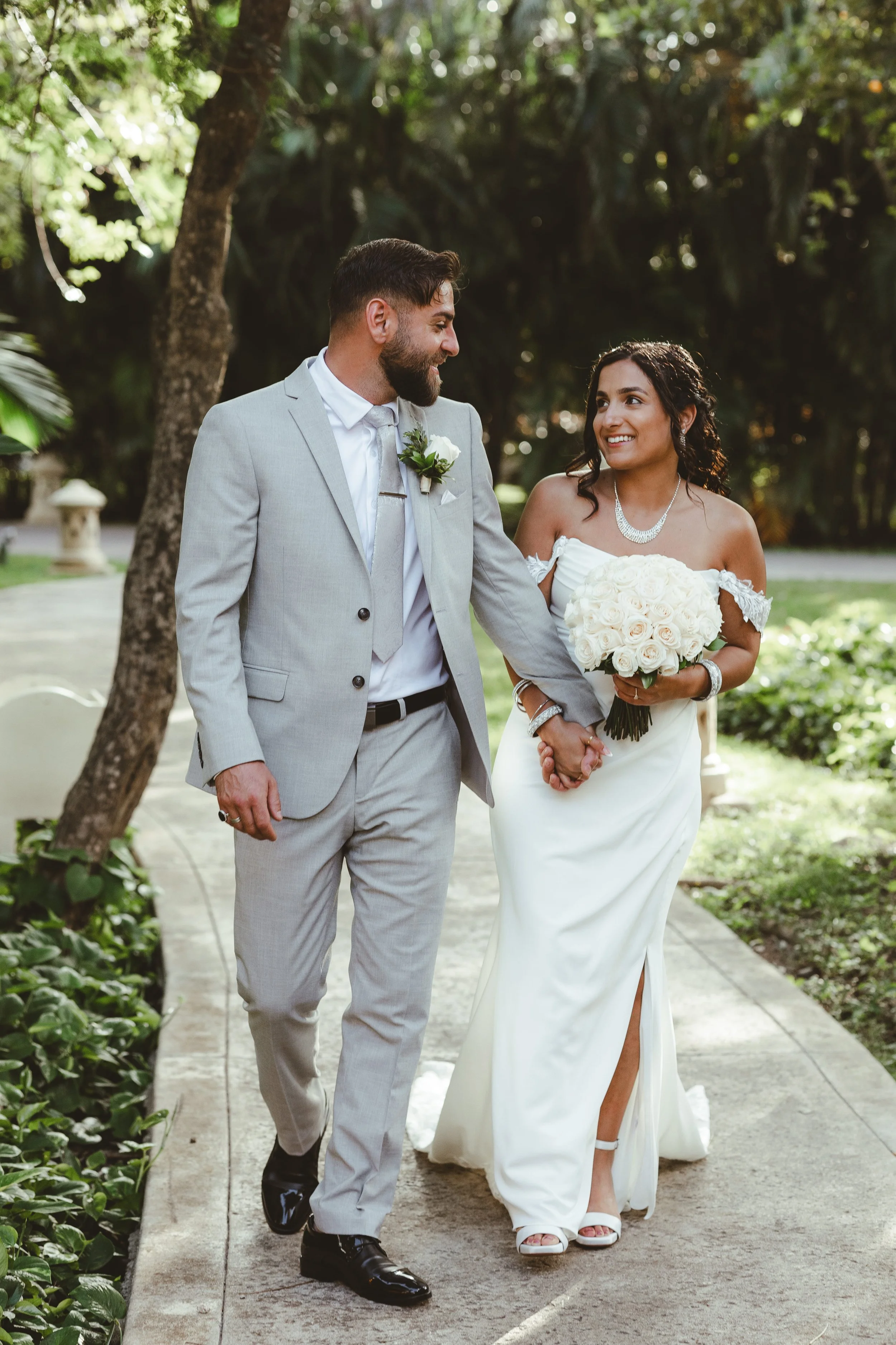 A newly married couple walking hand in hand outdoors, smiling at each other. The groom is wearing a light gray suit with a white shirt and black shoes, while the bride is in a white strapless wedding gown holding a bouquet of white roses. They are su