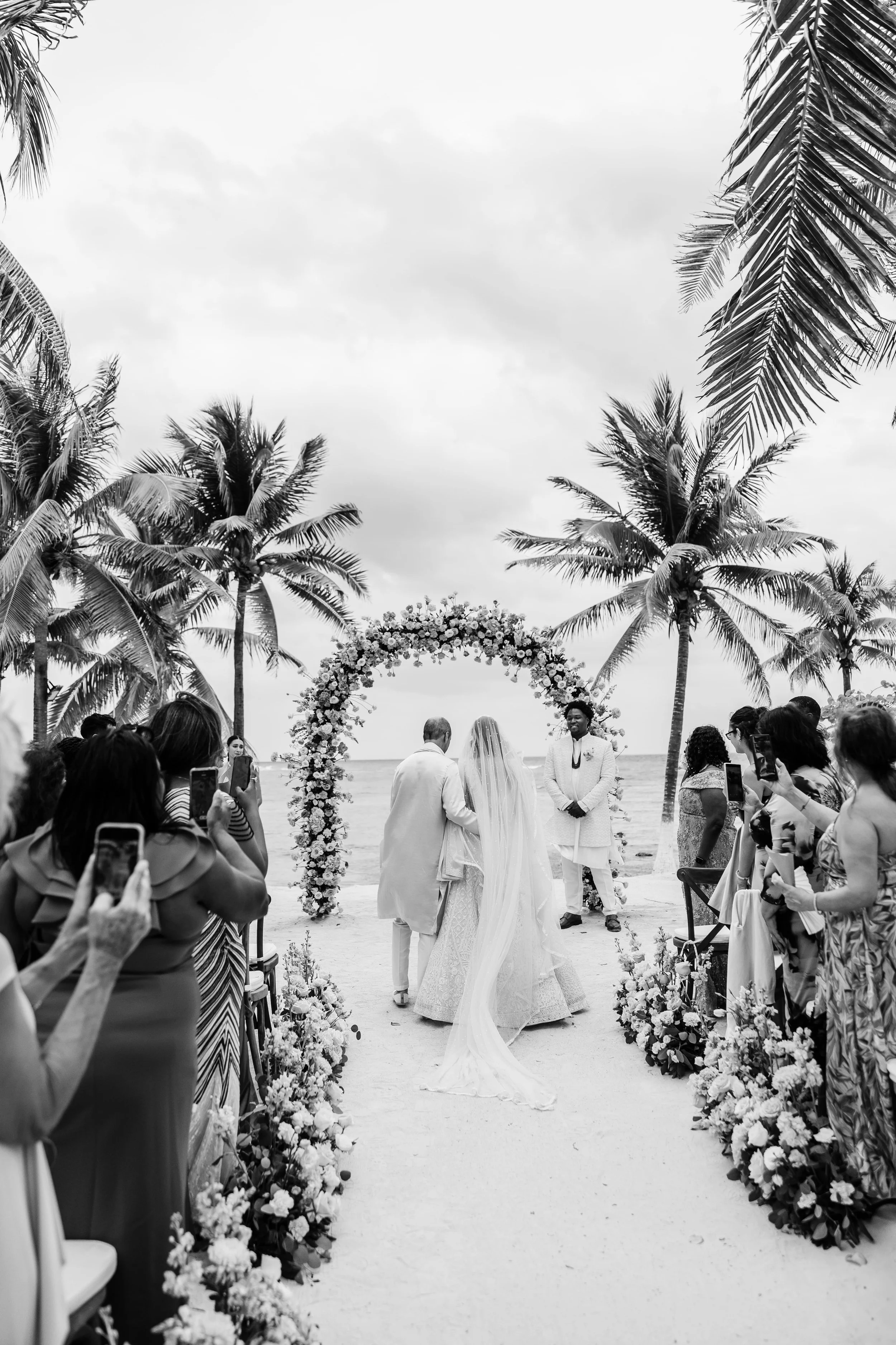 A black-and-white photo of a beach wedding ceremony with palm trees, an arch decorated with flowers, and people taking photos of the bride and groom.