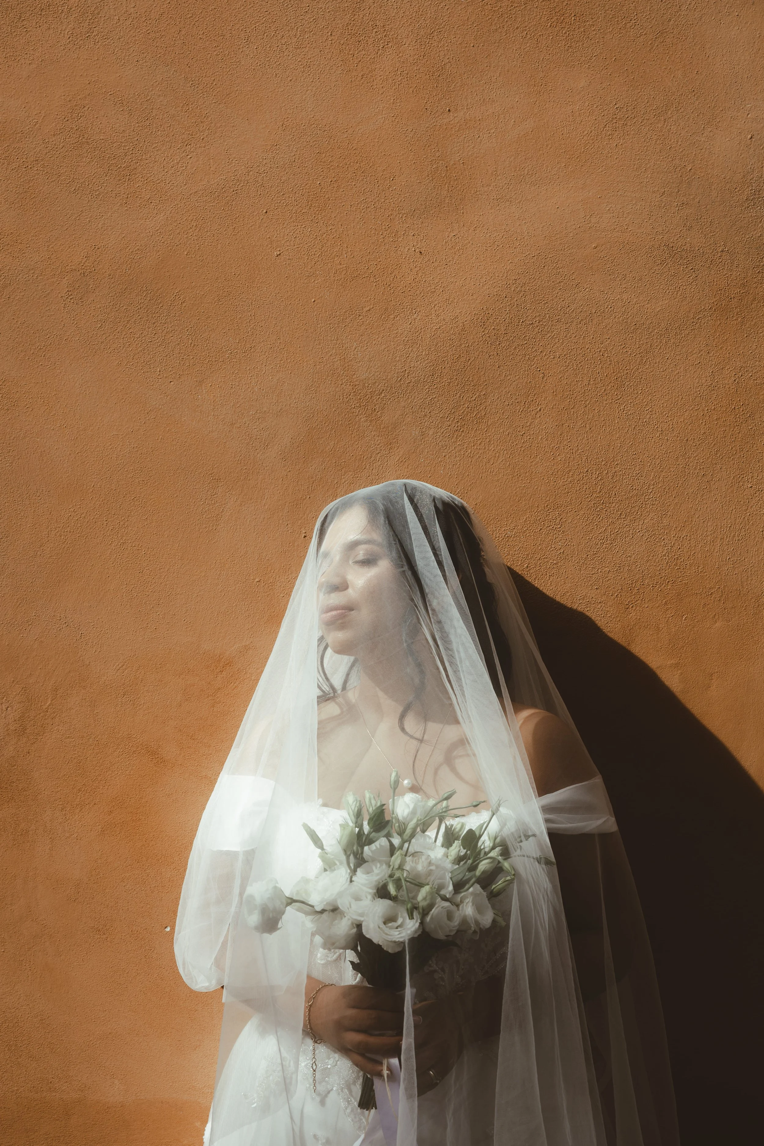 A woman dressed as a bride holding a bouquet of white flowers, with a sheer veil over her face, standing against an orange textured wall.