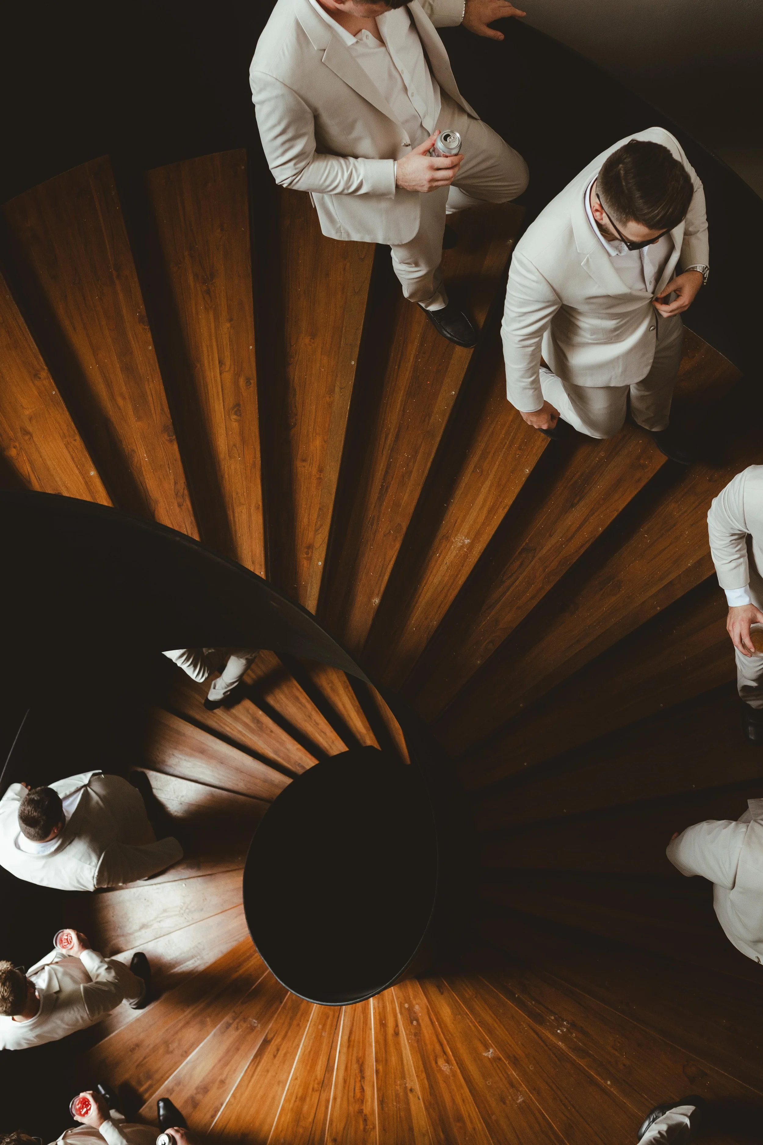 Overhead view of several groomsmen in light-colored suits standing on a wooden spiral staircase, holding drinks, seen from above, at Secrets Moxche Playa del Carmen.
