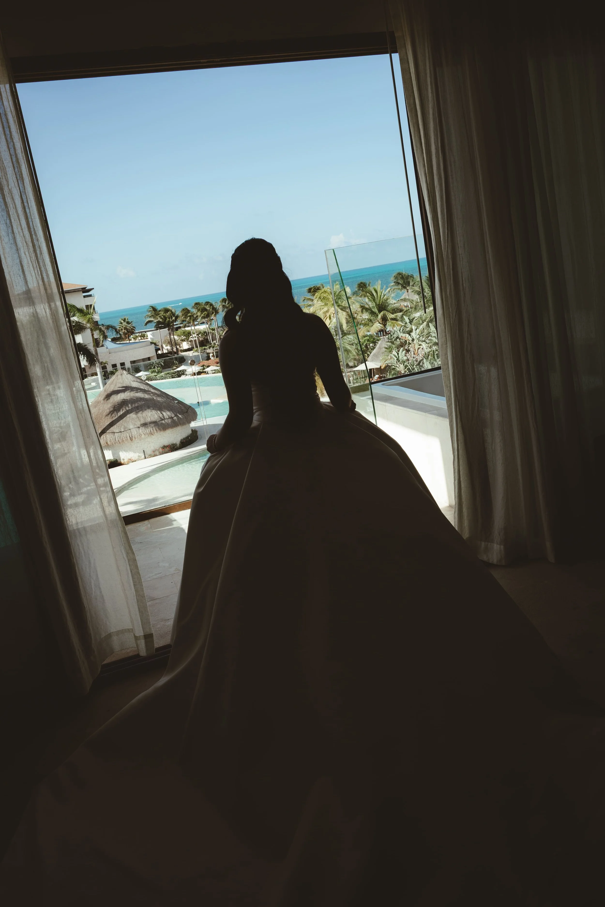 Silhouette of a person sitting by large window with ocean view, palm trees, and resort buildings in the background.