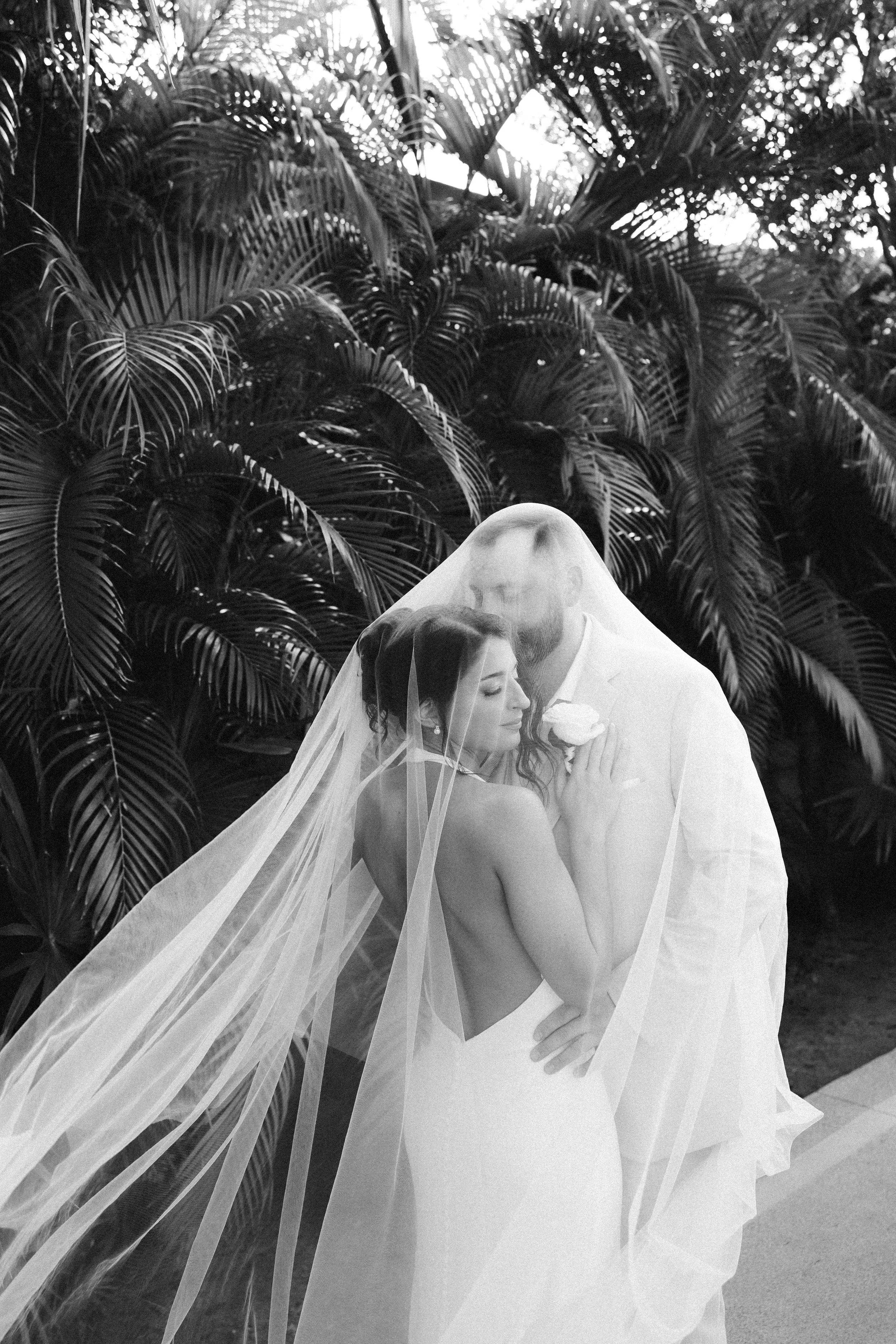 Black and white photo of a bride and groom embracing, with the bride wearing a wedding dress and veil, and the groom in a light-colored suit, standing in front of lush tropical plants.