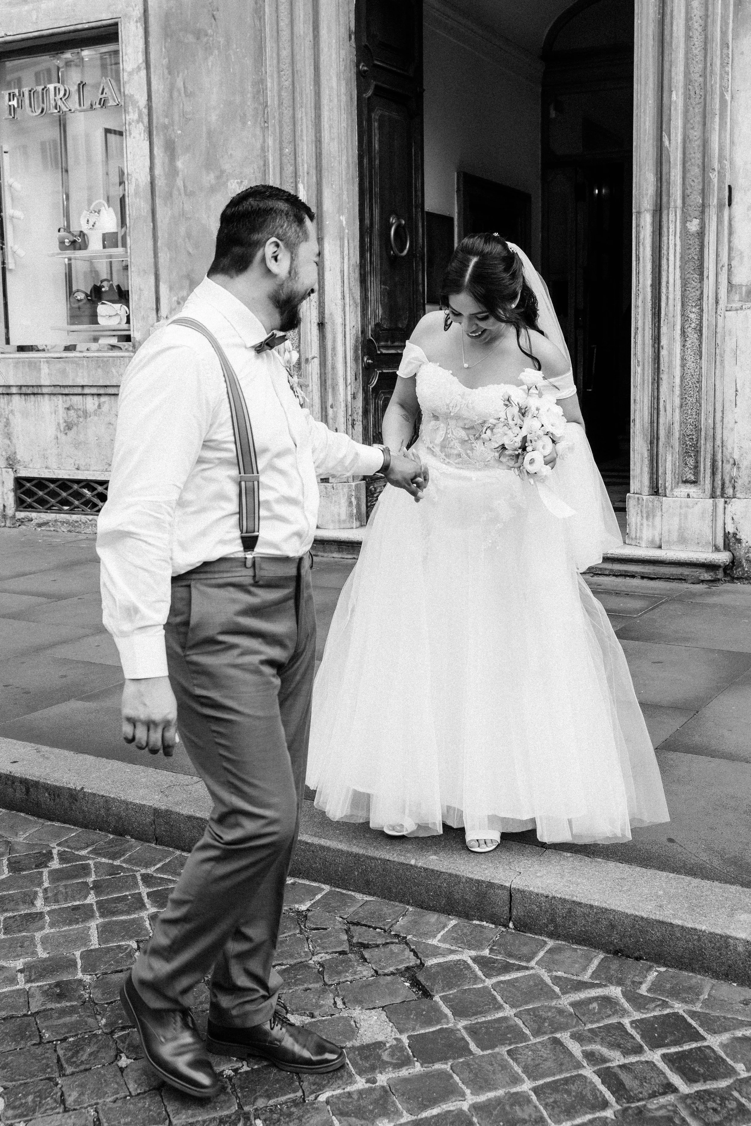 A black and white photo of a bride and groom outside a building. The groom, with dark hair and a beard, is holding the bride's hand and smiling. He is wearing dress pants, a shirt, suspenders, and a bow tie. The bride, with dark hair styled in waves,