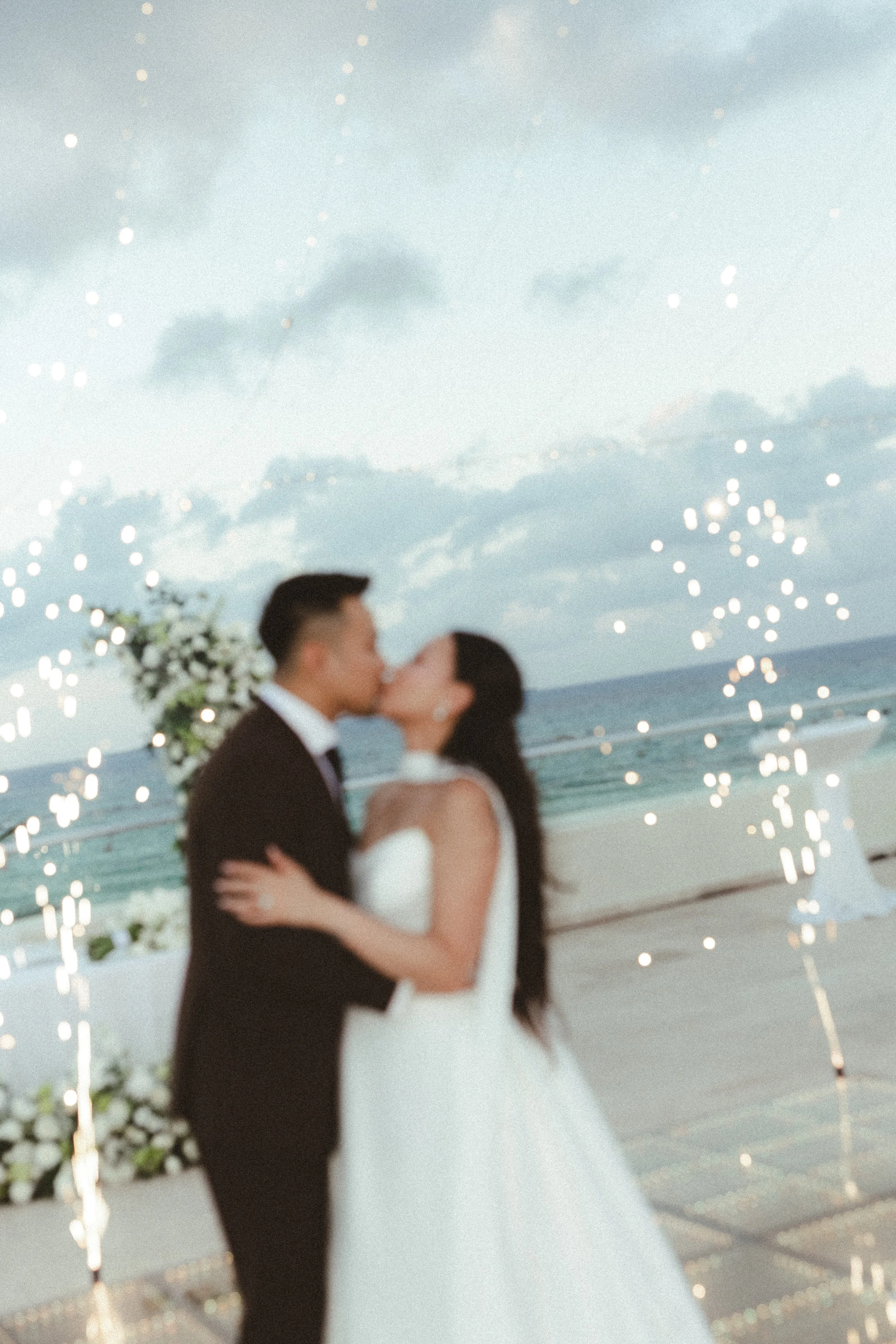 A couple dressed in wedding attire sharing a kiss on a beach during sunset or evening, with string lights overhead and the ocean in the background in Hyatt Ziva, Cancun.