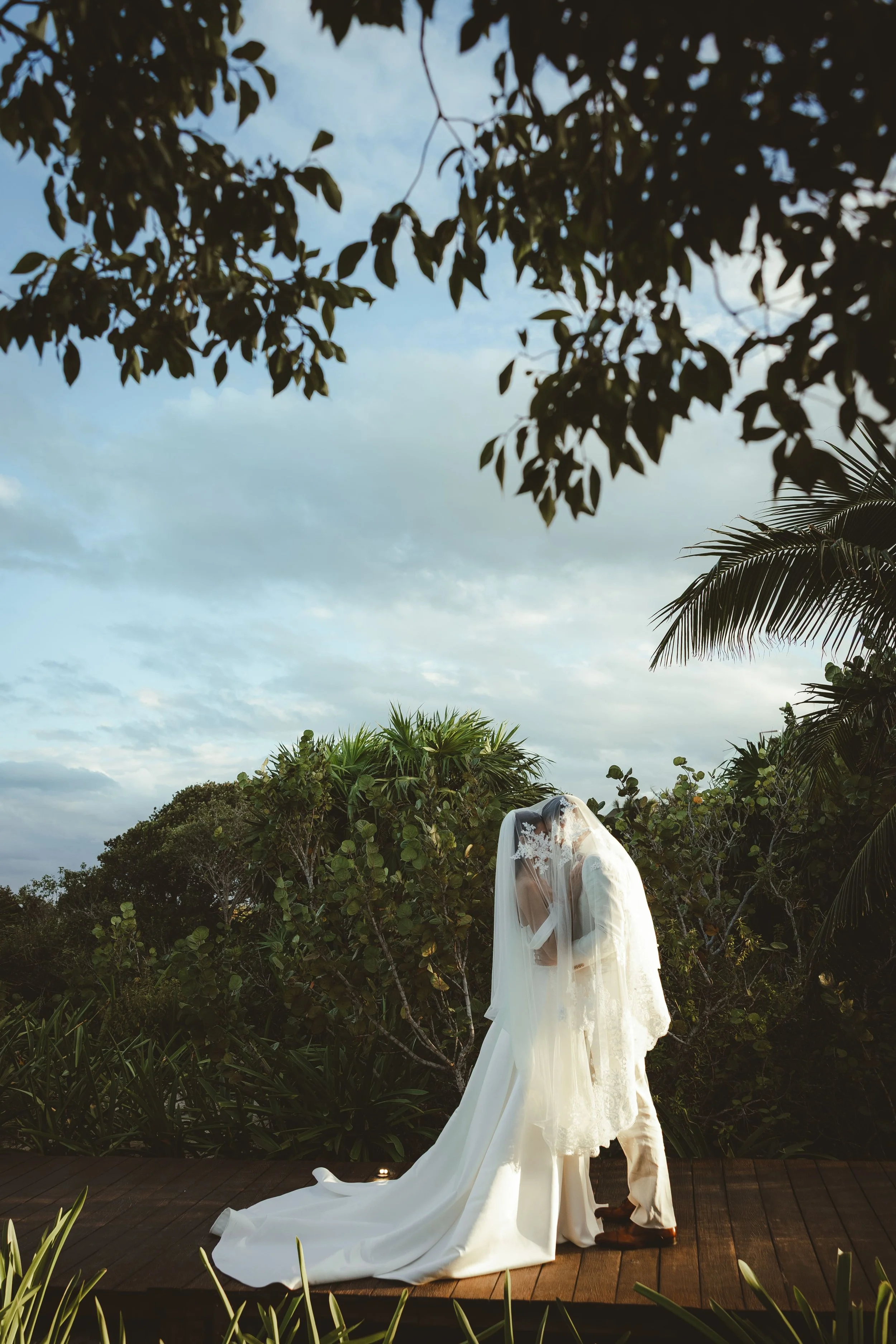 A bride in a white wedding dress with a long train and veil stands outdoors surrounded by greenery and trees during sunset, with some clouds in the sky.