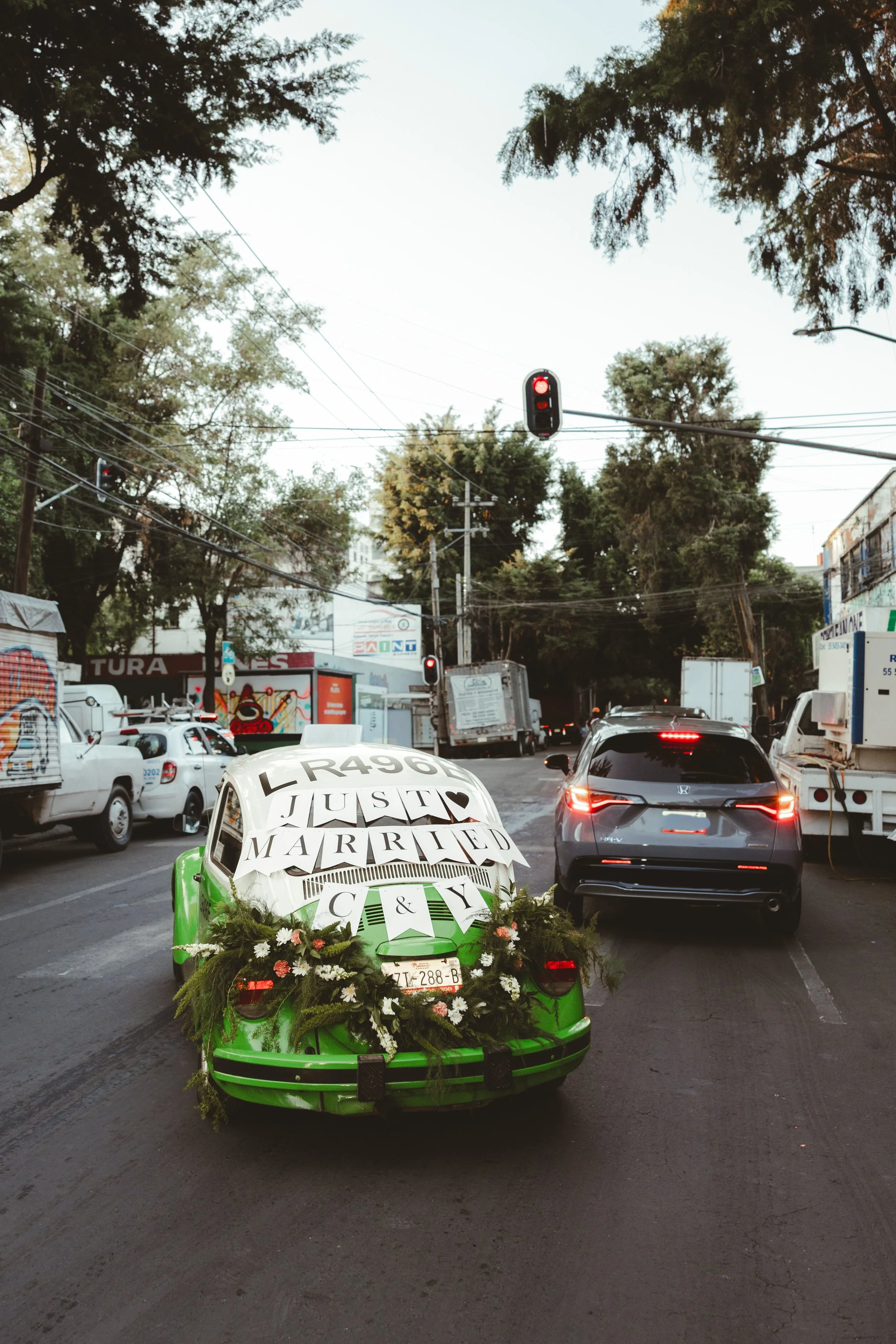 A green car decorated with flowers and wedding signs, including 'JUST MARRIED,' driving through a city street with traffic, trees, and buildings.
