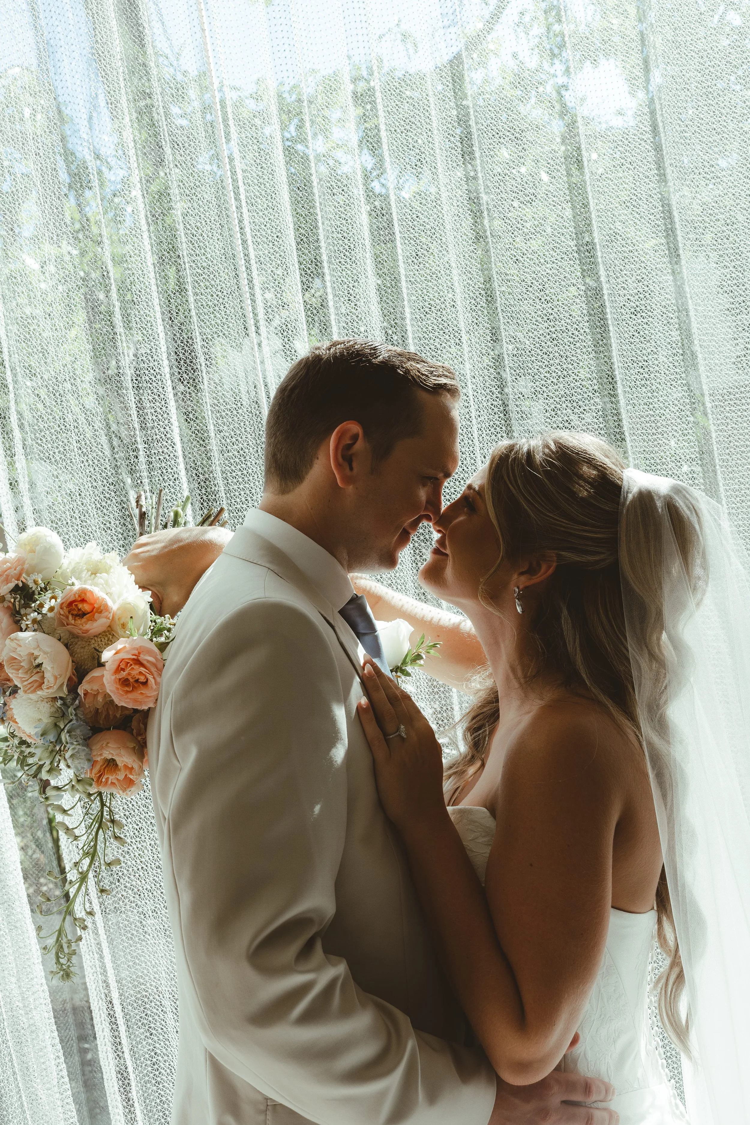 Bride and groom in a close embrace indoors with sheer curtains behind them, sunlight filtering through, flowers on the side.