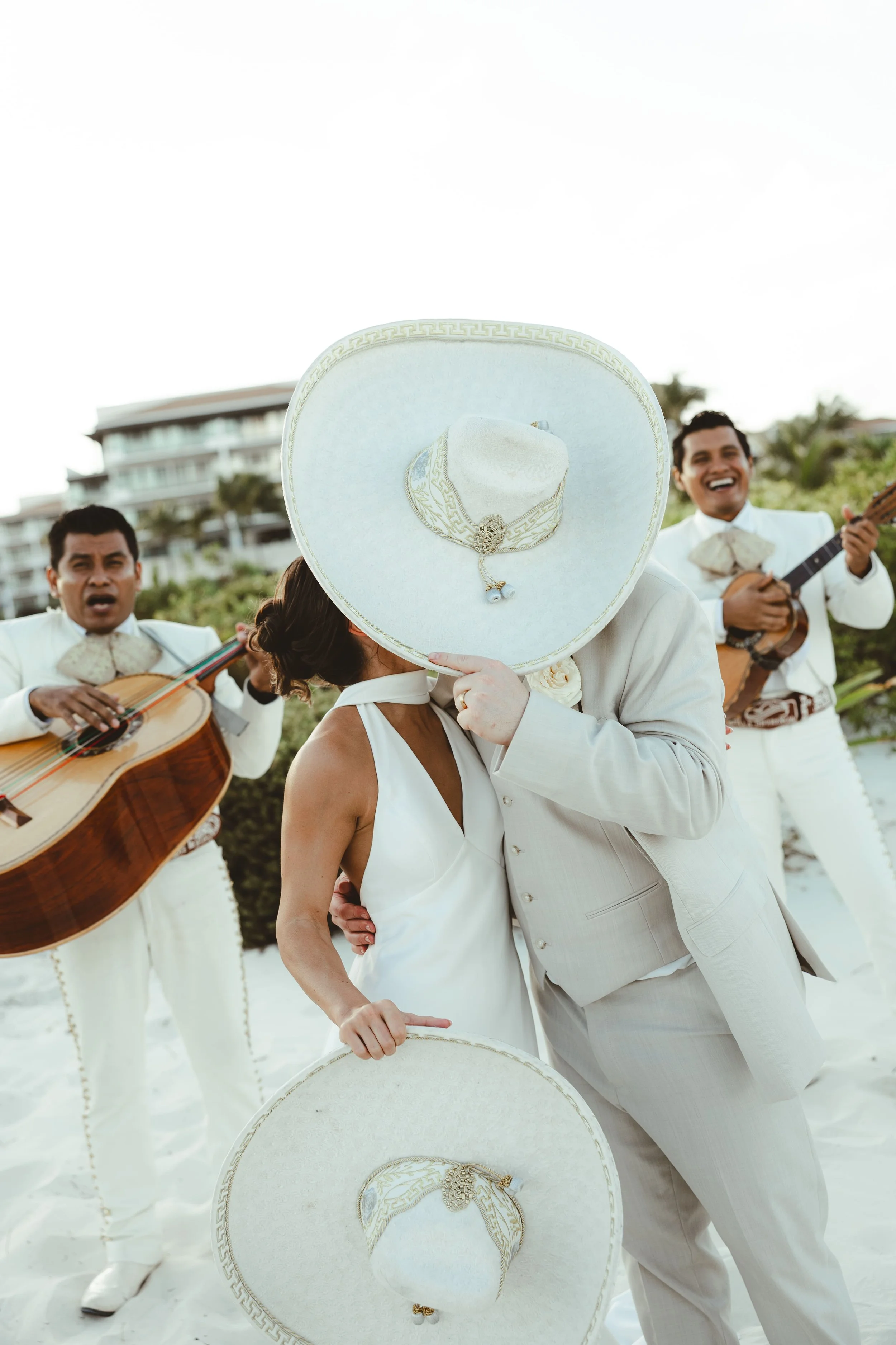 A couple dressed in white holding large white sombreros with gold trim during a daytime outdoor celebration, surrounded by musicians playing guitars.