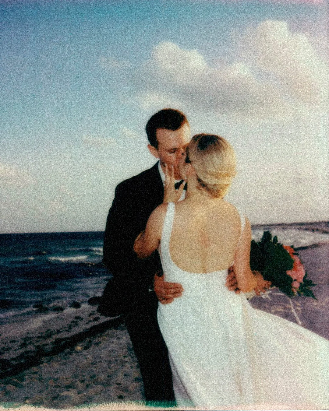 A couple dressed in wedding attire kissing on the beach during sunset, with the ocean and sky in the background.