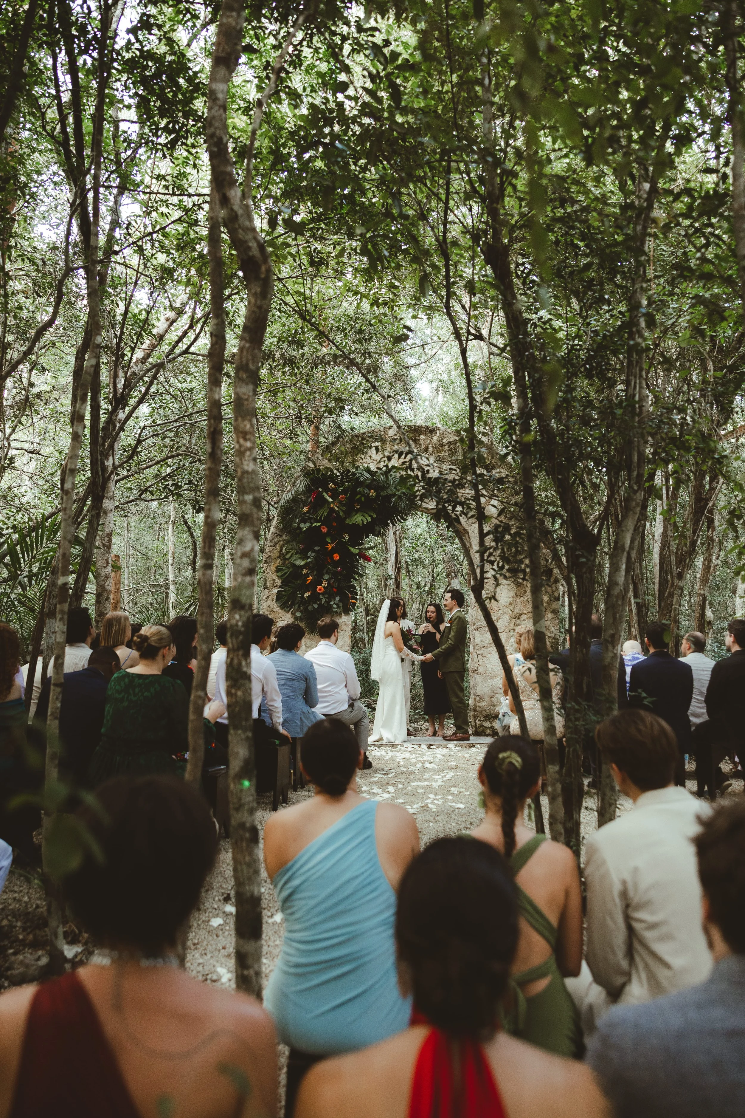 A wedding ceremony taking place outdoors under trees with guests seated on either side, and the bride and groom standing together as they exchange vows in front of a floral arch.
