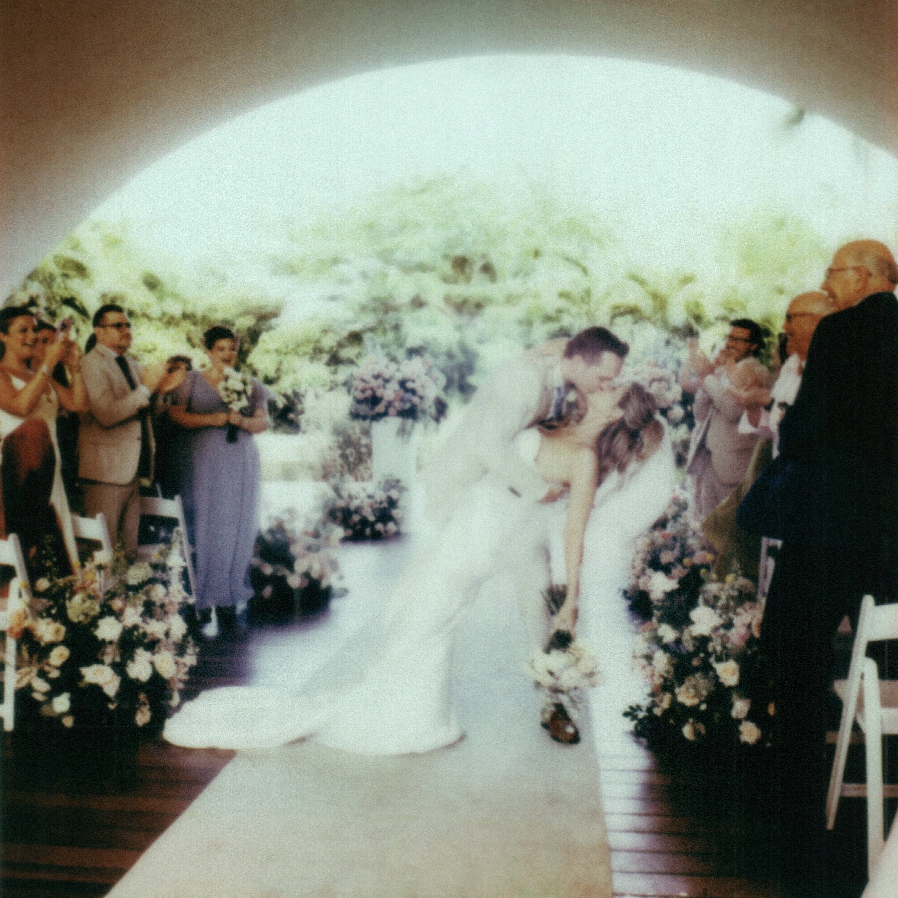 A bride and groom standing under a floral archway at their wedding, surrounded by guests taking pictures and smiling.