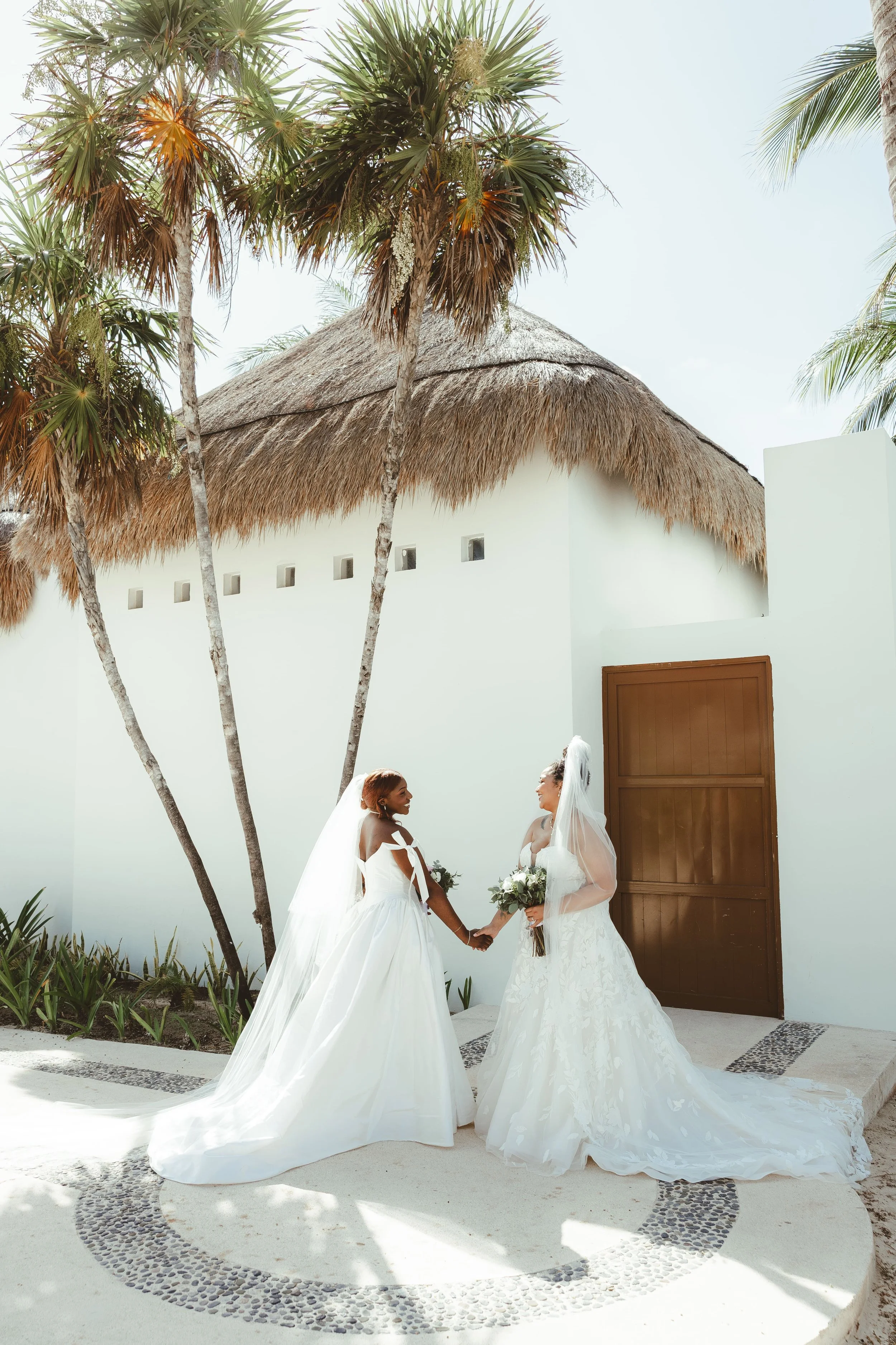 Two brides in white wedding dresses holding hands and smiling at each other, standing outdoors near a white wall with a thatched roof. There are trees and plants around, and the ground has a circular pebble design.