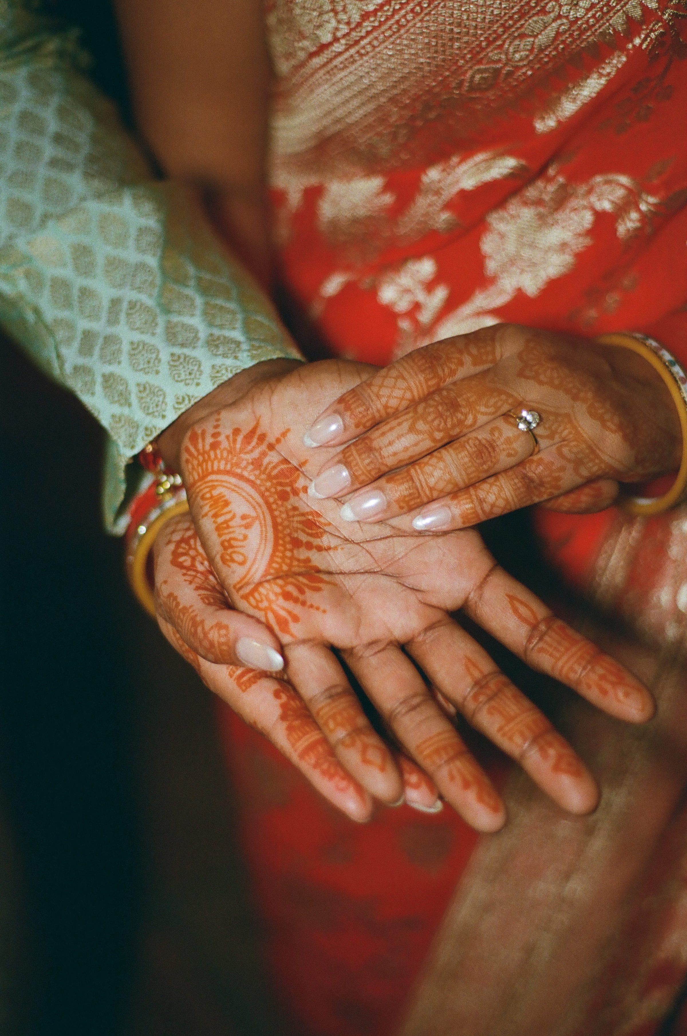 Close-up of a person's hands with intricate henna designs, jewelry, and a wedding ring, resting on their lap covered in red and gold traditional attire.