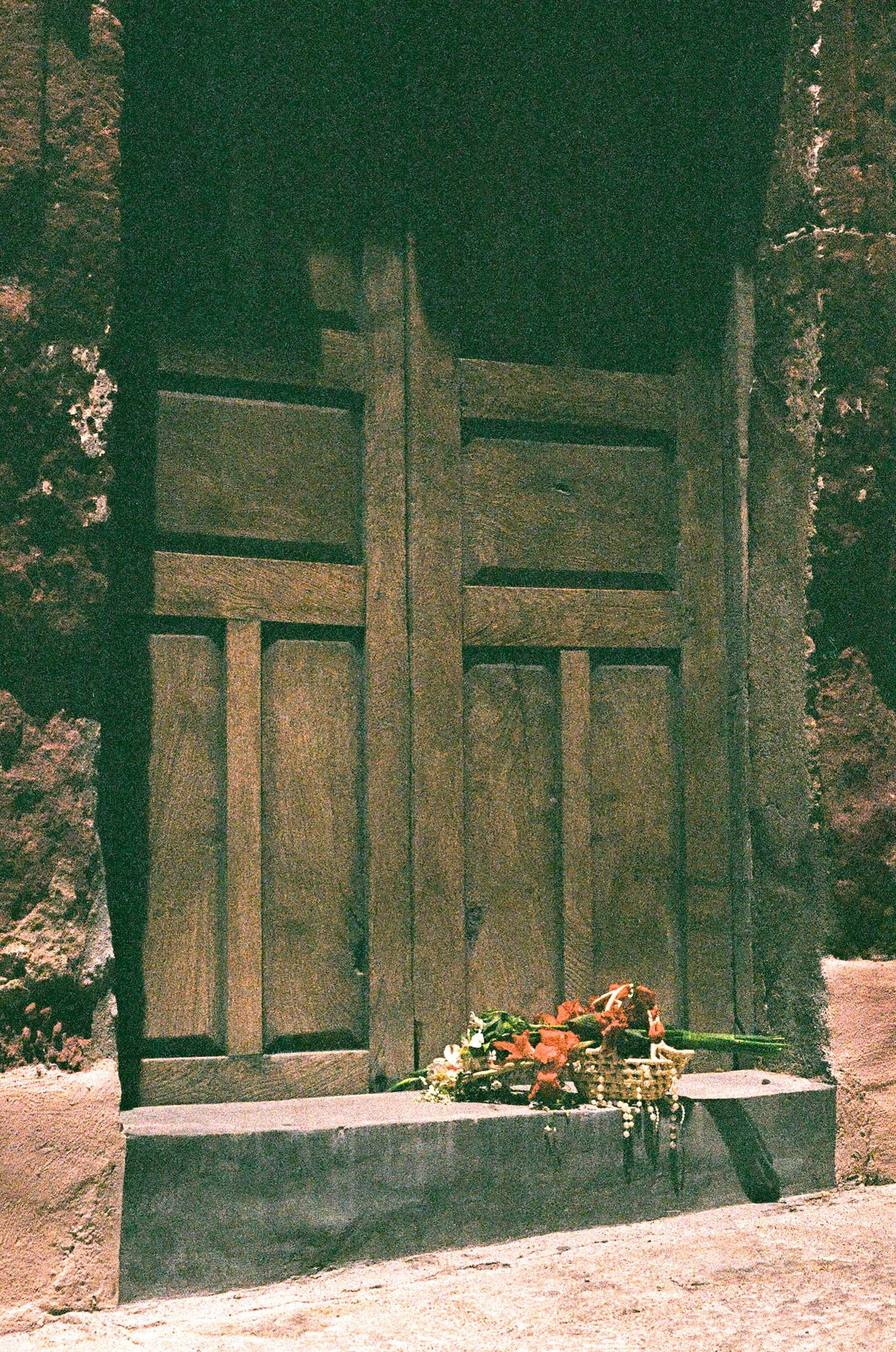 A wooden door with decorative panels, located in a stone wall. There is a flower arrangement and rosary beads on a concrete ledge in front of the door.