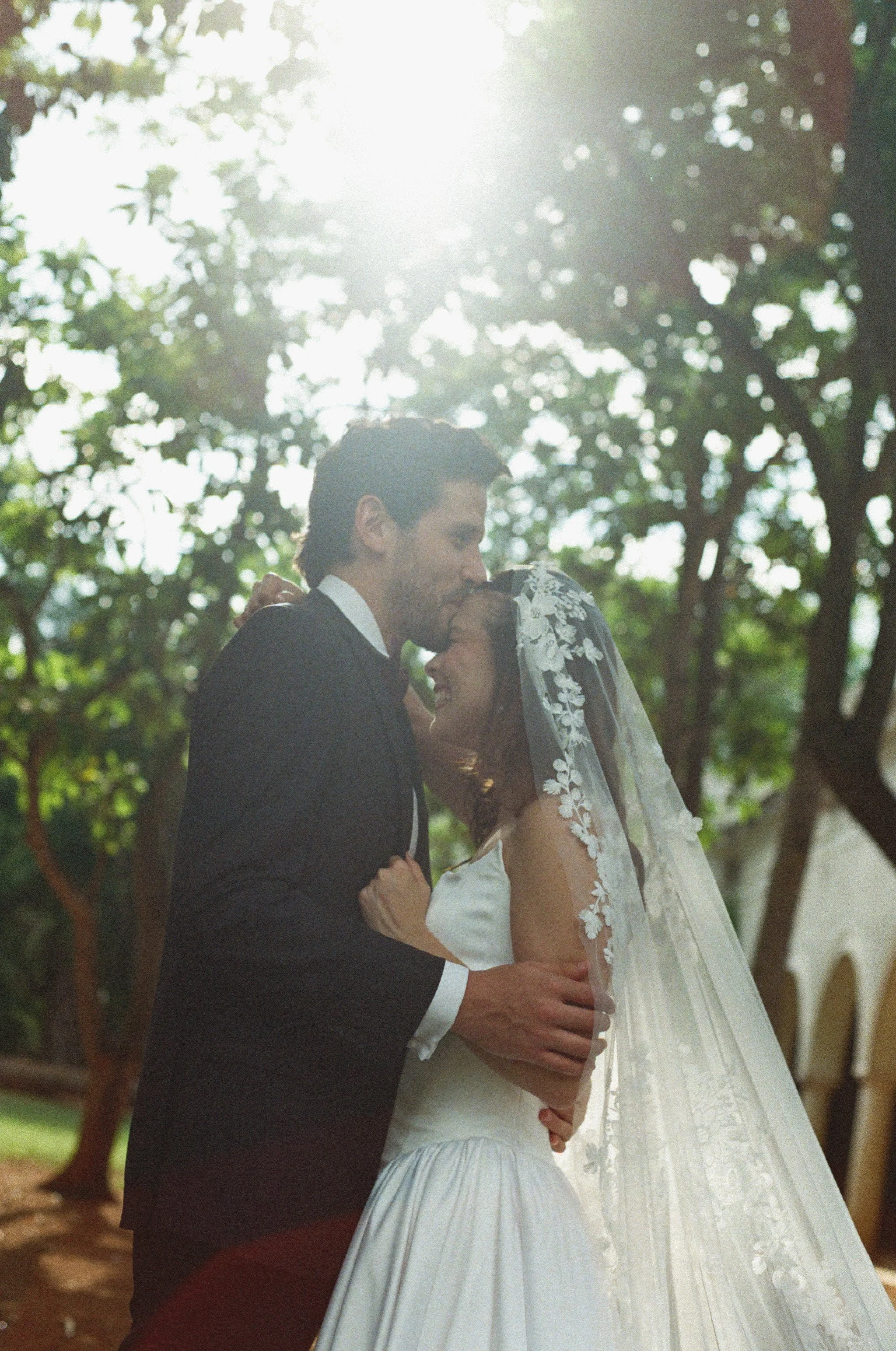 A bride and groom sharing a kiss outdoors during daytime with sunlight filtering through trees in the background.