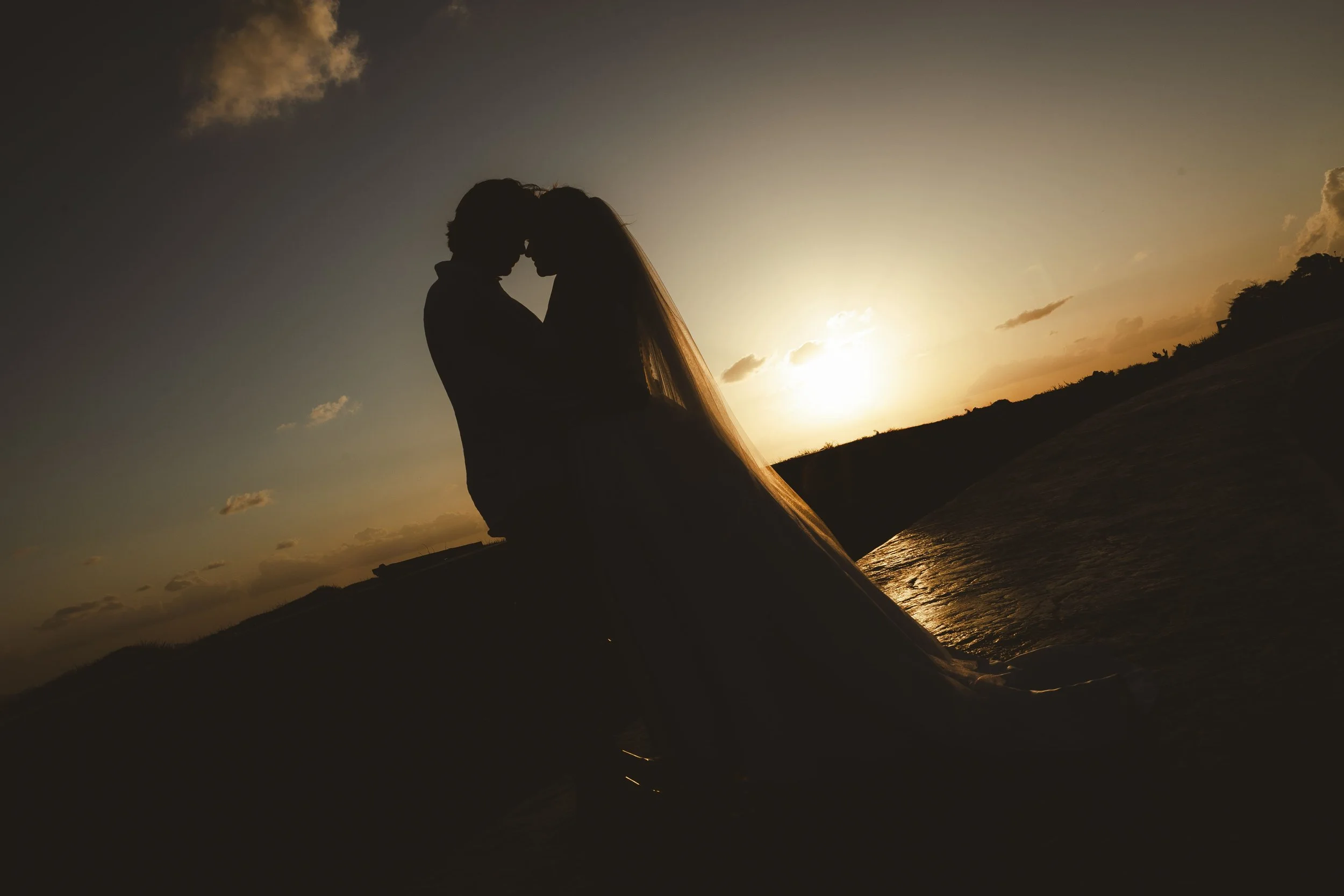 Silhouettes of a couple embracing on a mountain, with the sun setting behind them, creating a warm glow in the sky.