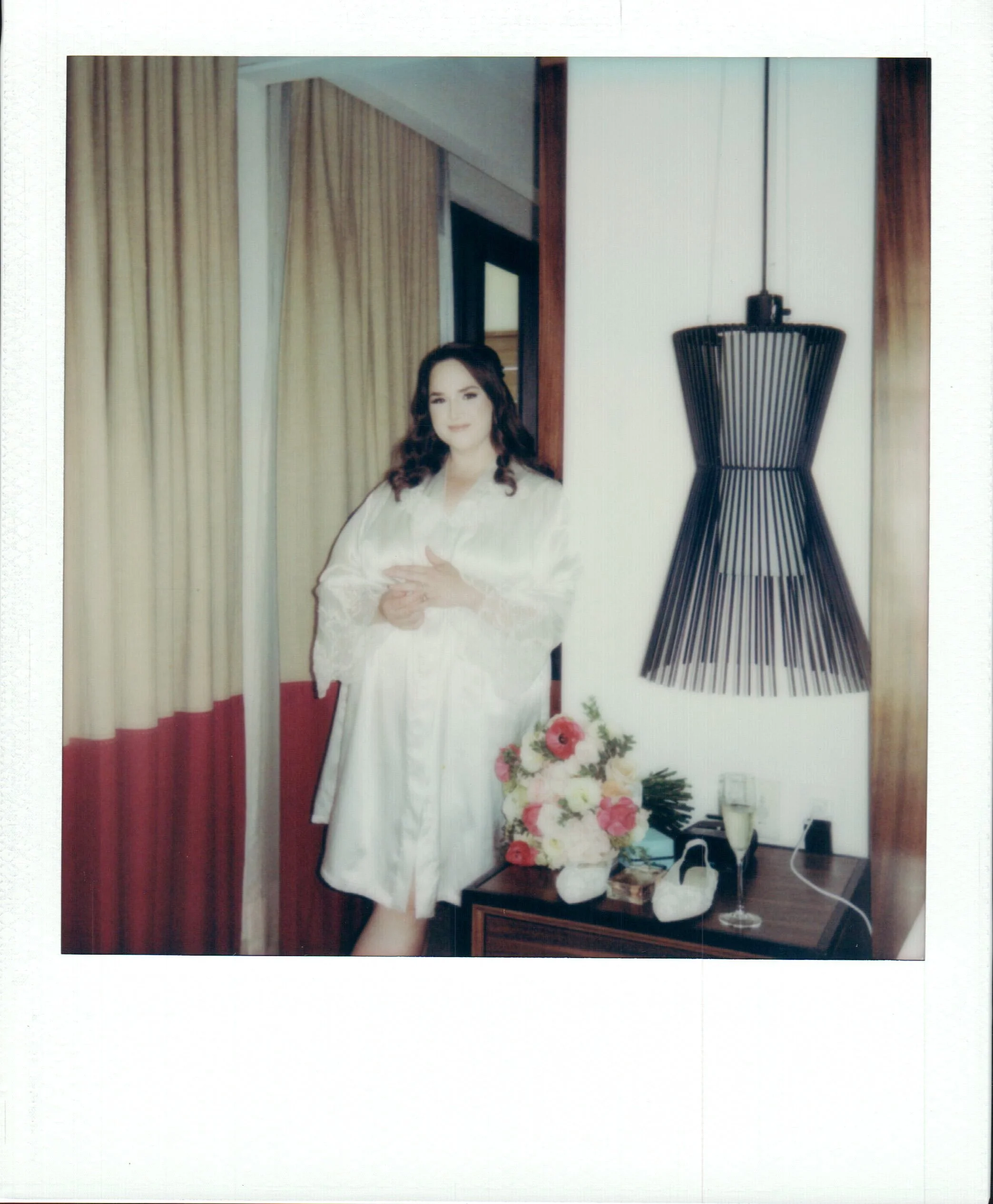 A woman in a white robe standing next to a bouquet of flowers on a table, with curtains in the background and a large black decorative light fixture on the wall.