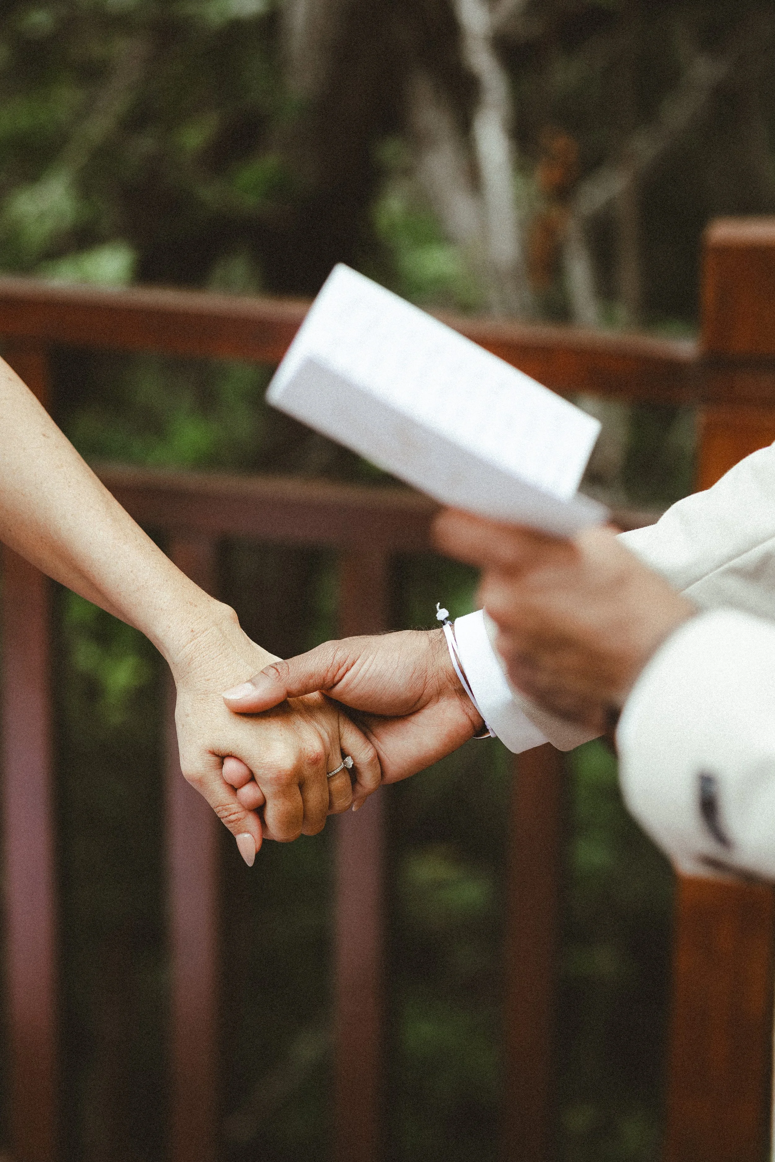 A person in a white shirt is holding hands with another individual, with a blurred background of greenery and a fence.