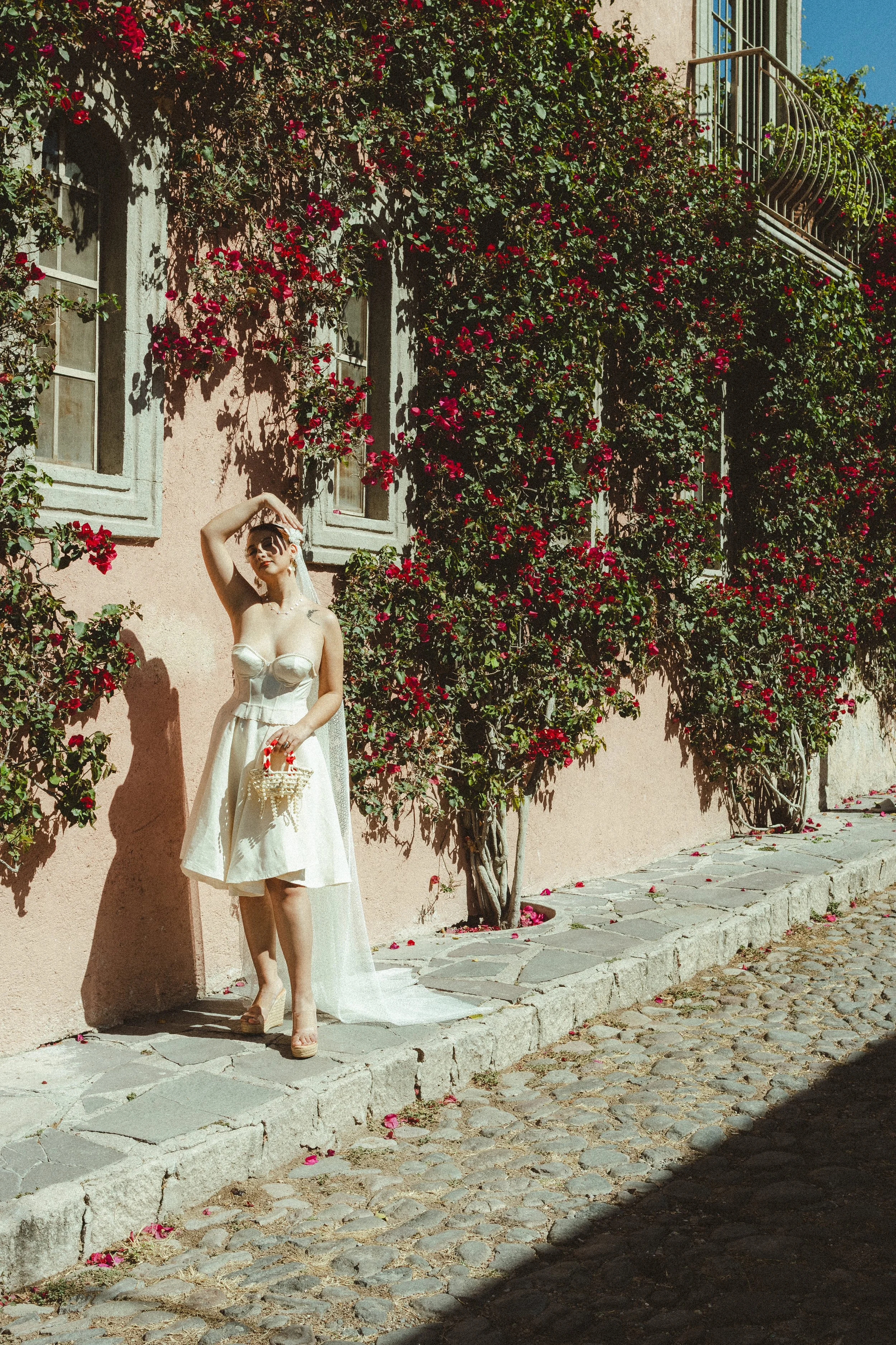 A woman in a white strapless dress standing on a cobblestone street beside a pink stucco building covered in pink flowers. The woman holds a small basket and has her right hand raised above her head, shading her eyes from the sun. The building has se