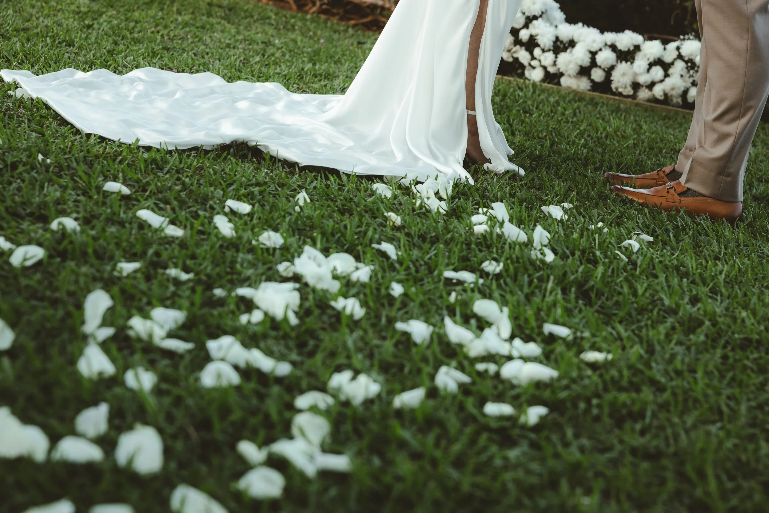 A bride and groom standing on grass with white rose petals scattered around, the bride wearing a white satin wedding dress, and the groom in tan trousers and brown shoes.