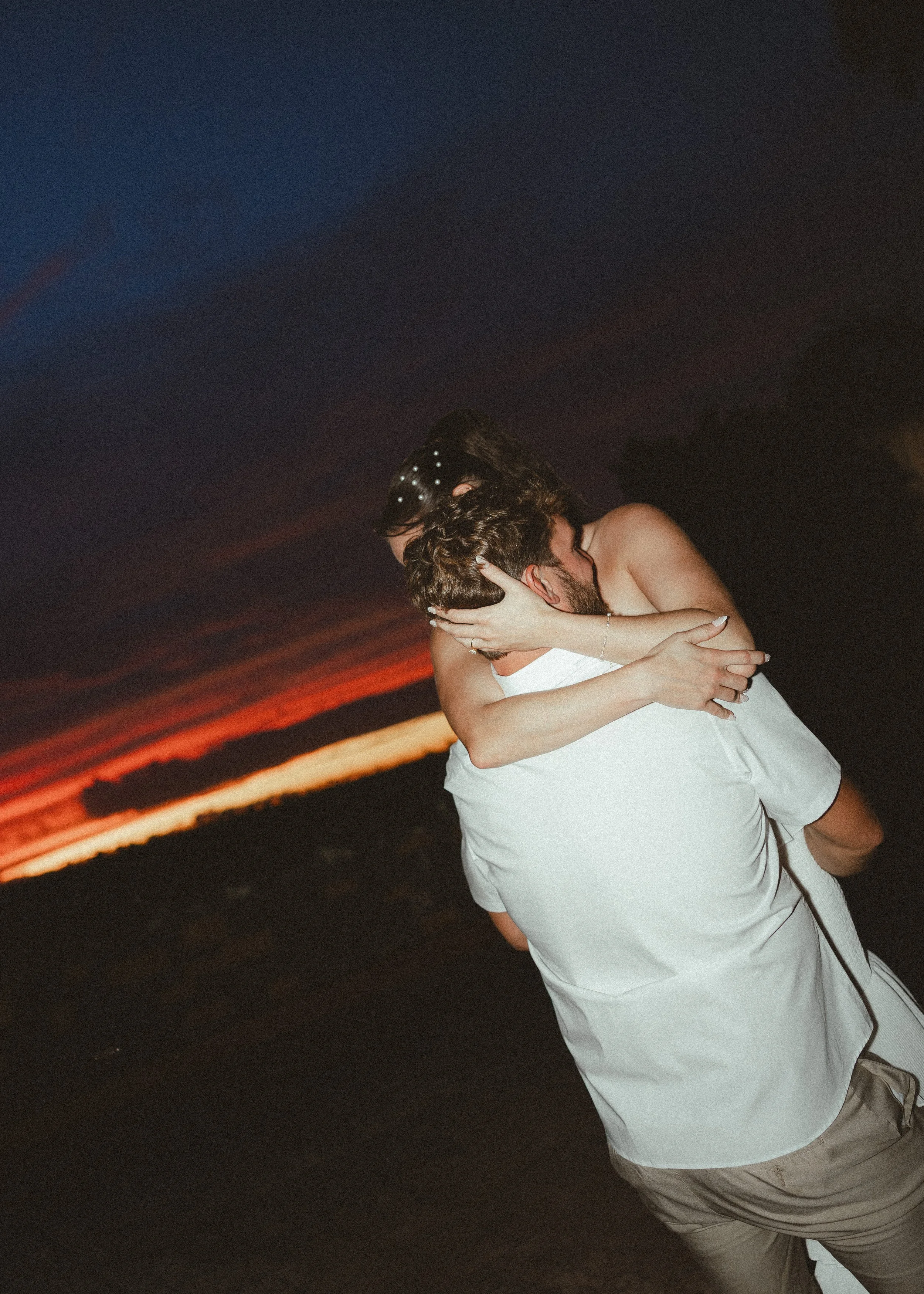 A couple hugging outdoors during sunset, with a dark sky and orange horizon in the background.