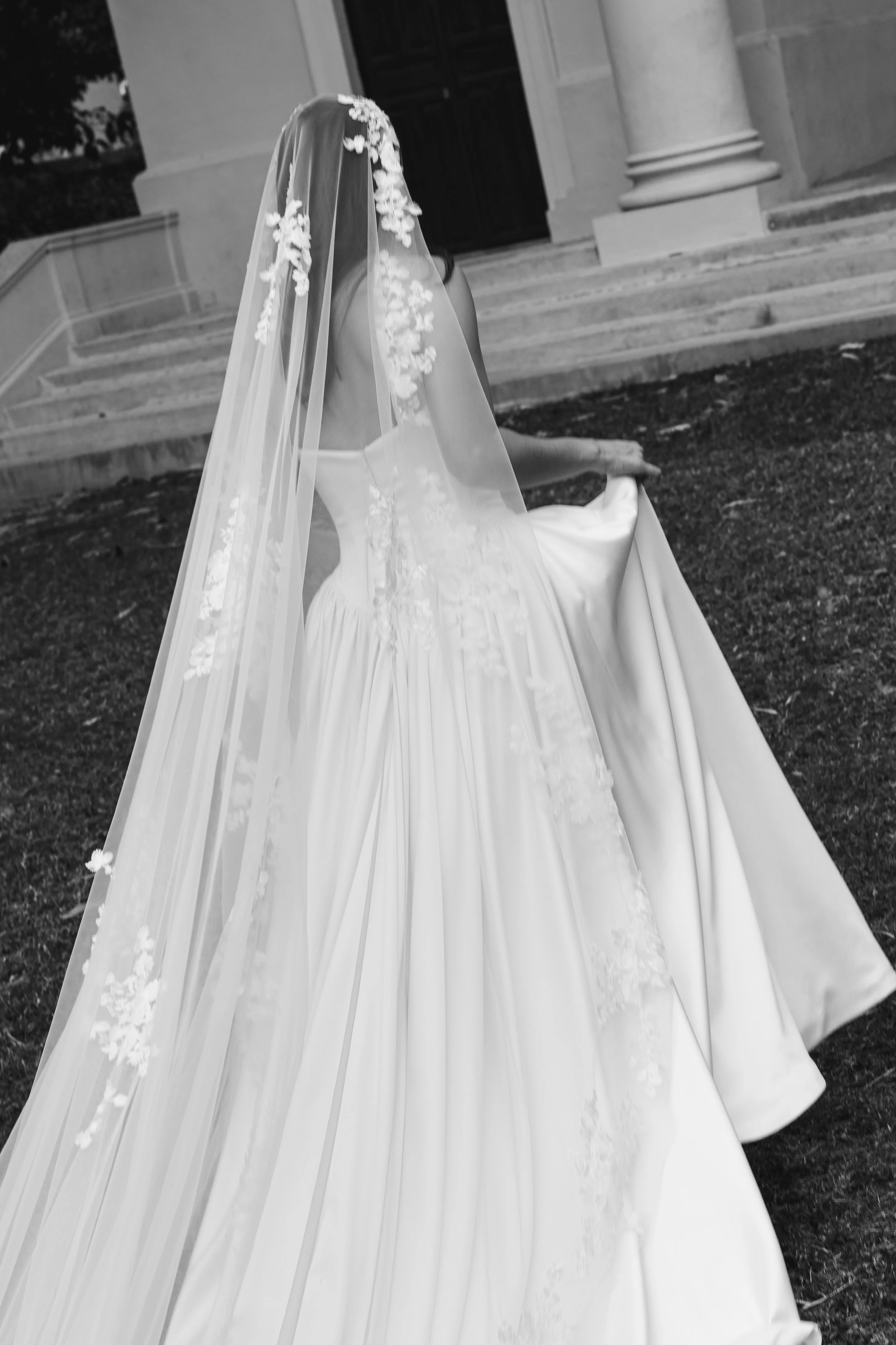 A woman dressed in a wedding gown and veil holding the train of her wedding dress, standing outside in front of a building with pillars and stairs in black and white photo.