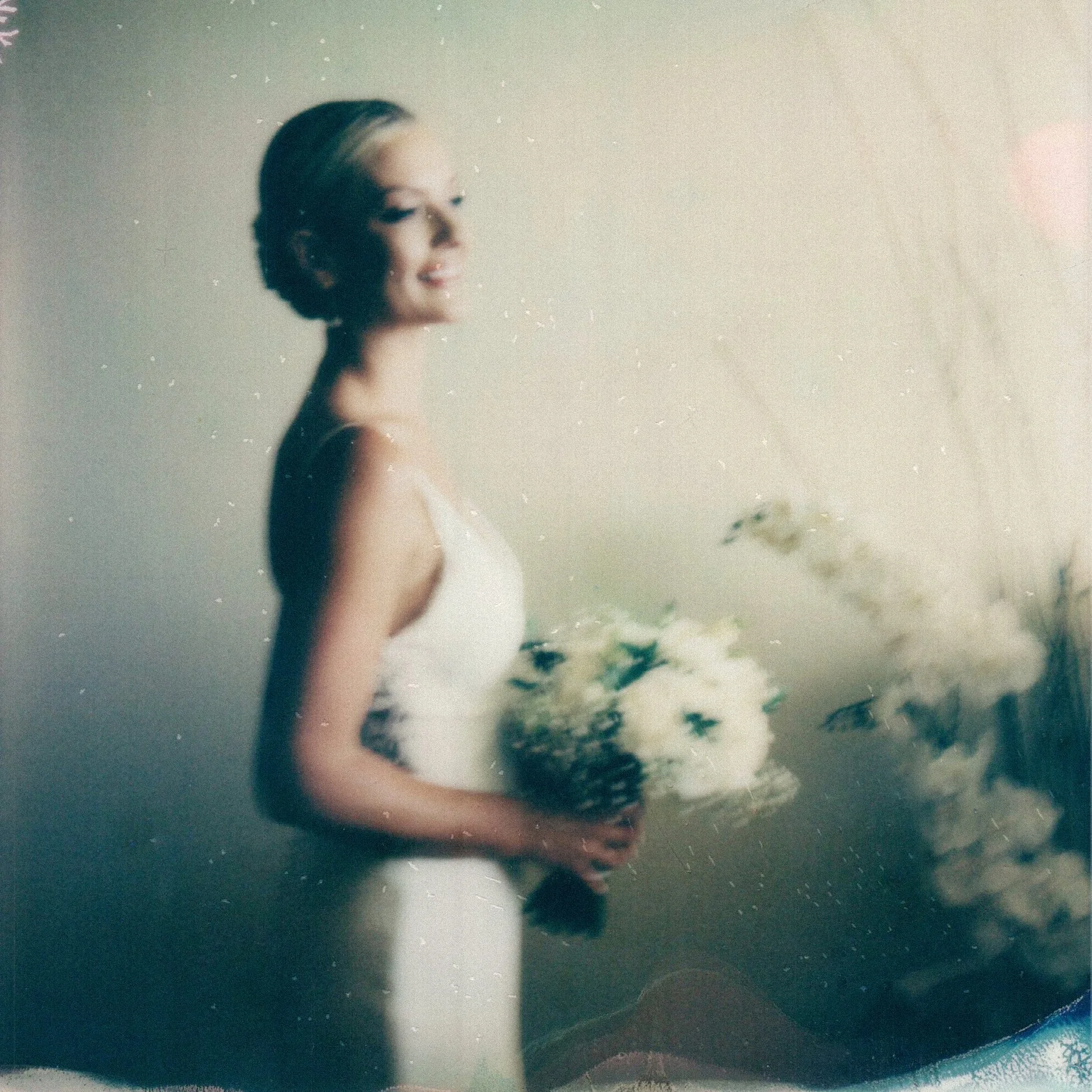 A bride with short blonde hair in a bun, wearing a white dress, smiling and looking to the side, reflected in a mirror with a foggy, textured surface. Some flowers are visible in the background.