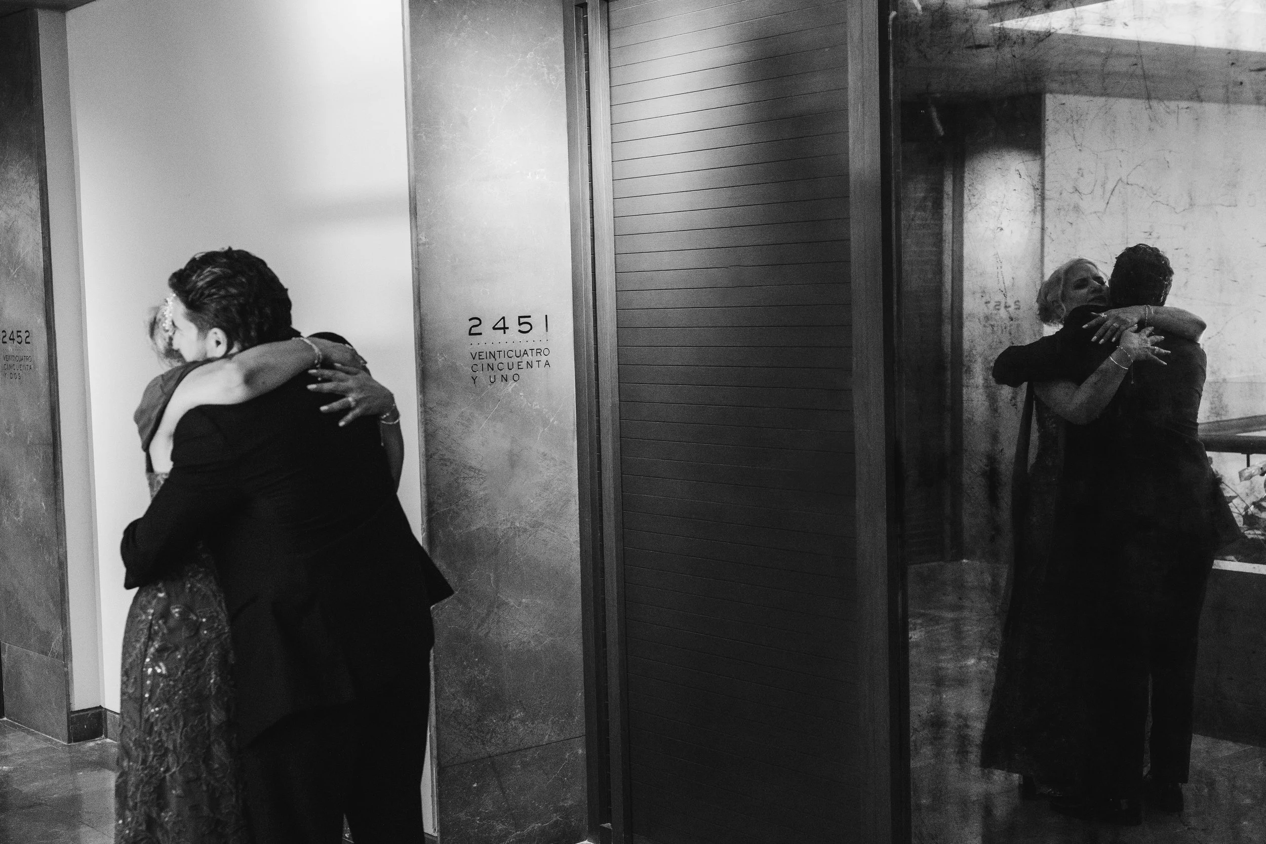 Two women hugging in front of elevator doors. One woman's back facing camera, the other's face visible and smiling.