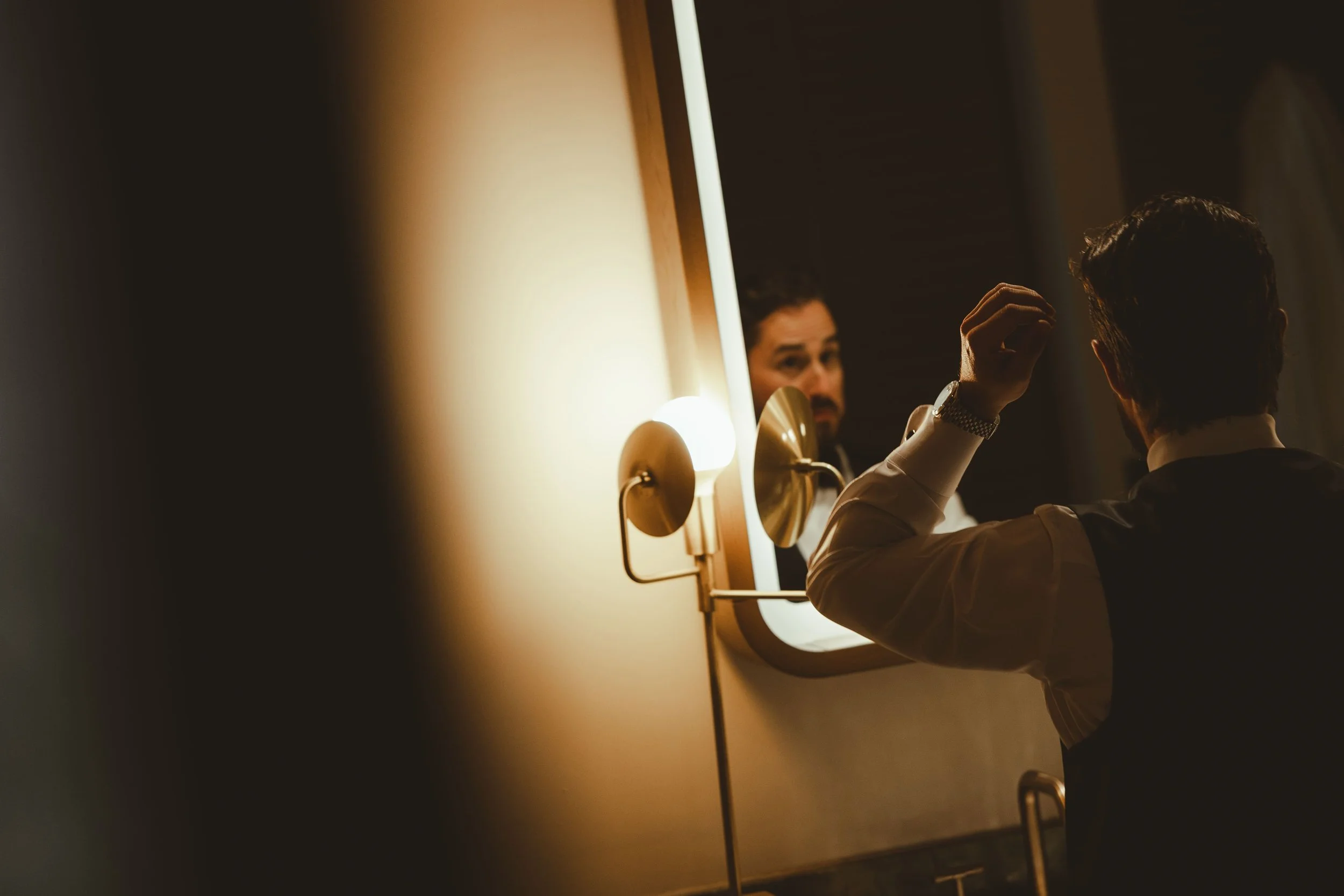 A man looking at himself in a mirror while styling his hair in a dimly lit room with warm lighting.