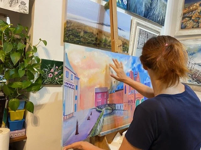 Miriam de Nardi sta dipingendo un paesaggio urbano con edifici colorati, un ponte e un cielo al tramonto