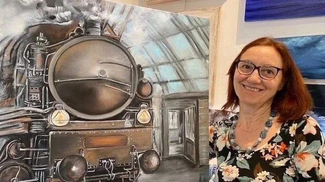 Donna sorridente con occhiali e vestito floreale posizionata accanto a un dipinto di una locomotiva a vapore.