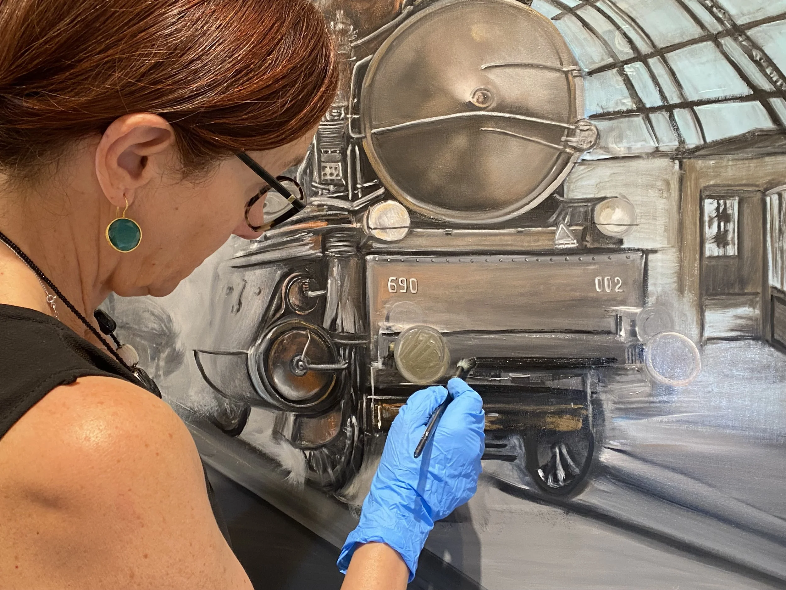 Miriam De Nardi sta dipingendo un quadro di un treno a olio, visibile a destra dell'immagine, indossando guanti blu.