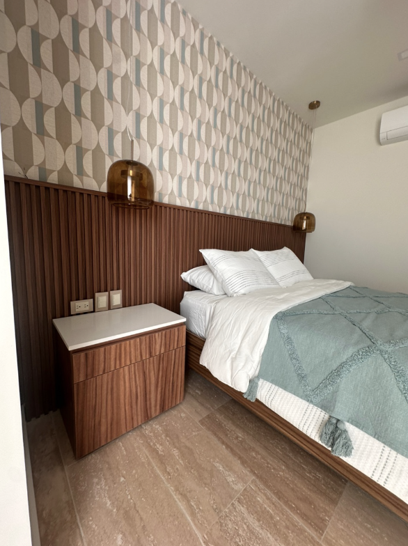 Habitación moderna con cama doble, cabecera de madera, paredes con papel tapiz decorativo y lámparas colgantes de color ámbar, con mesa de noche de madera y pisos de piedra