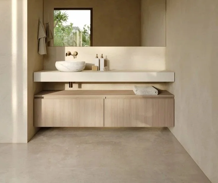 Lavabo de piedra sobre encimera blanca en un baño minimalista con espejo grande, ventana pequeña y toallas dobladas sobre un mueble de madera debajo.