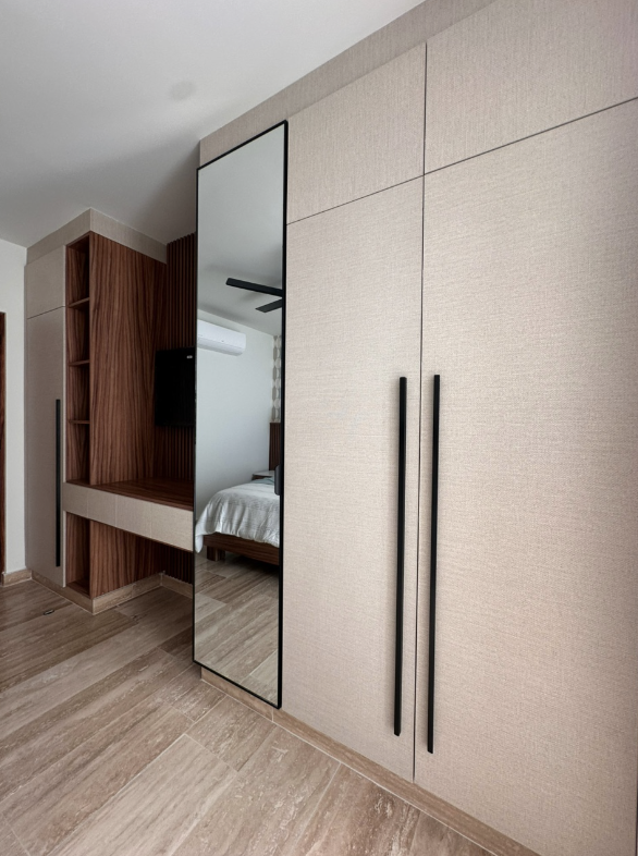 Armario grande con puertas beige y tiradores largos negros, espejo empotrado en una de las puertas refleja una cama y una pared con aires acondicionados, y estantería de madera junto a la pared
