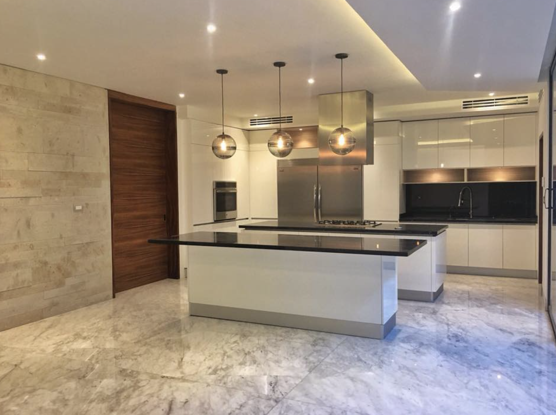 Cocina moderna con isla central, gabinetes blancos, pared de piedra, luces colgantes, electrodomésticos de acero inoxidable y piso de mármol.