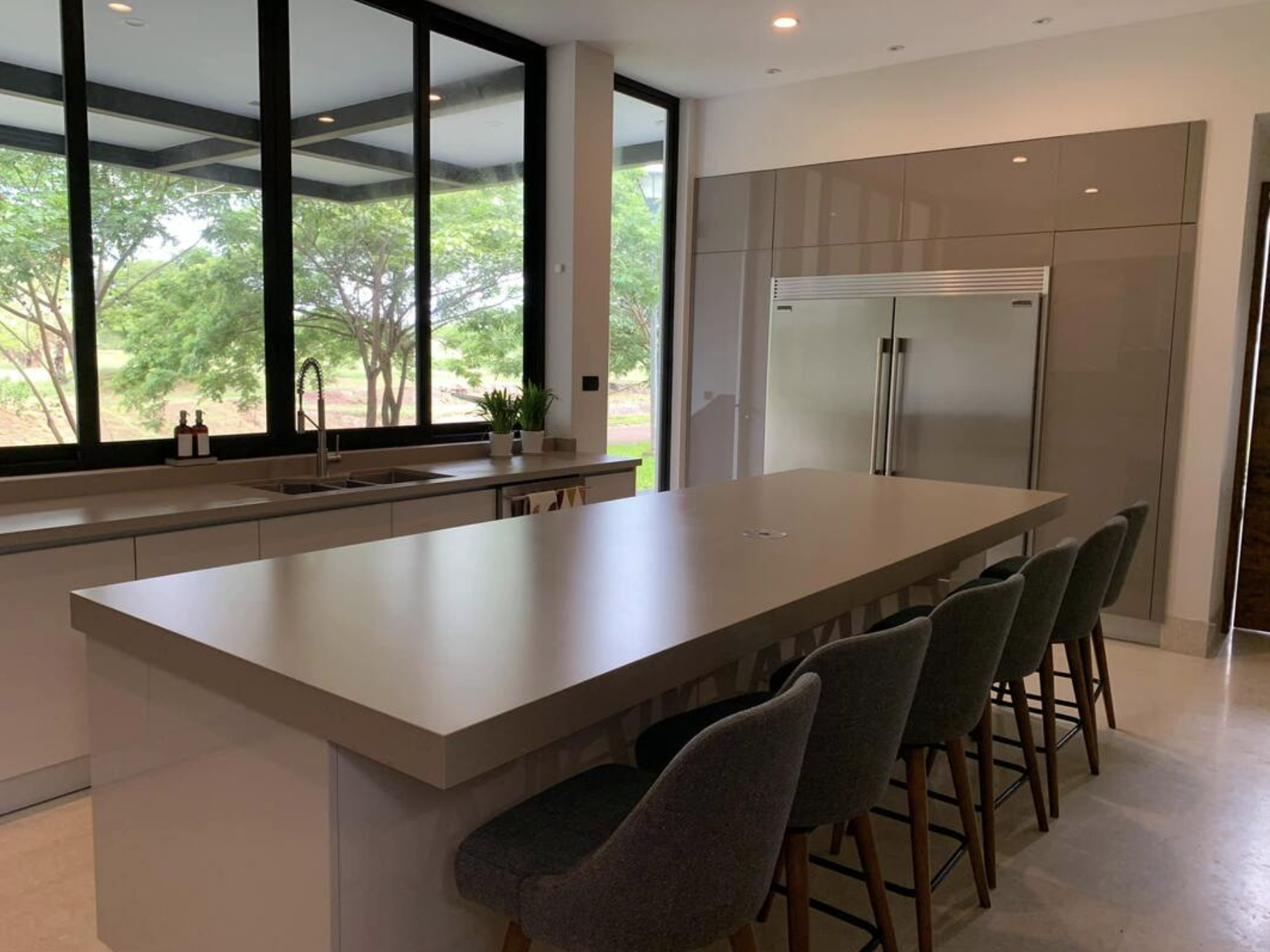 Cocina moderna con barra y sillas, grandes ventanas que dejan ver árboles verdes, refrigerador de acero inoxidable, plantas en la mesa, interiores claros y luminosos.